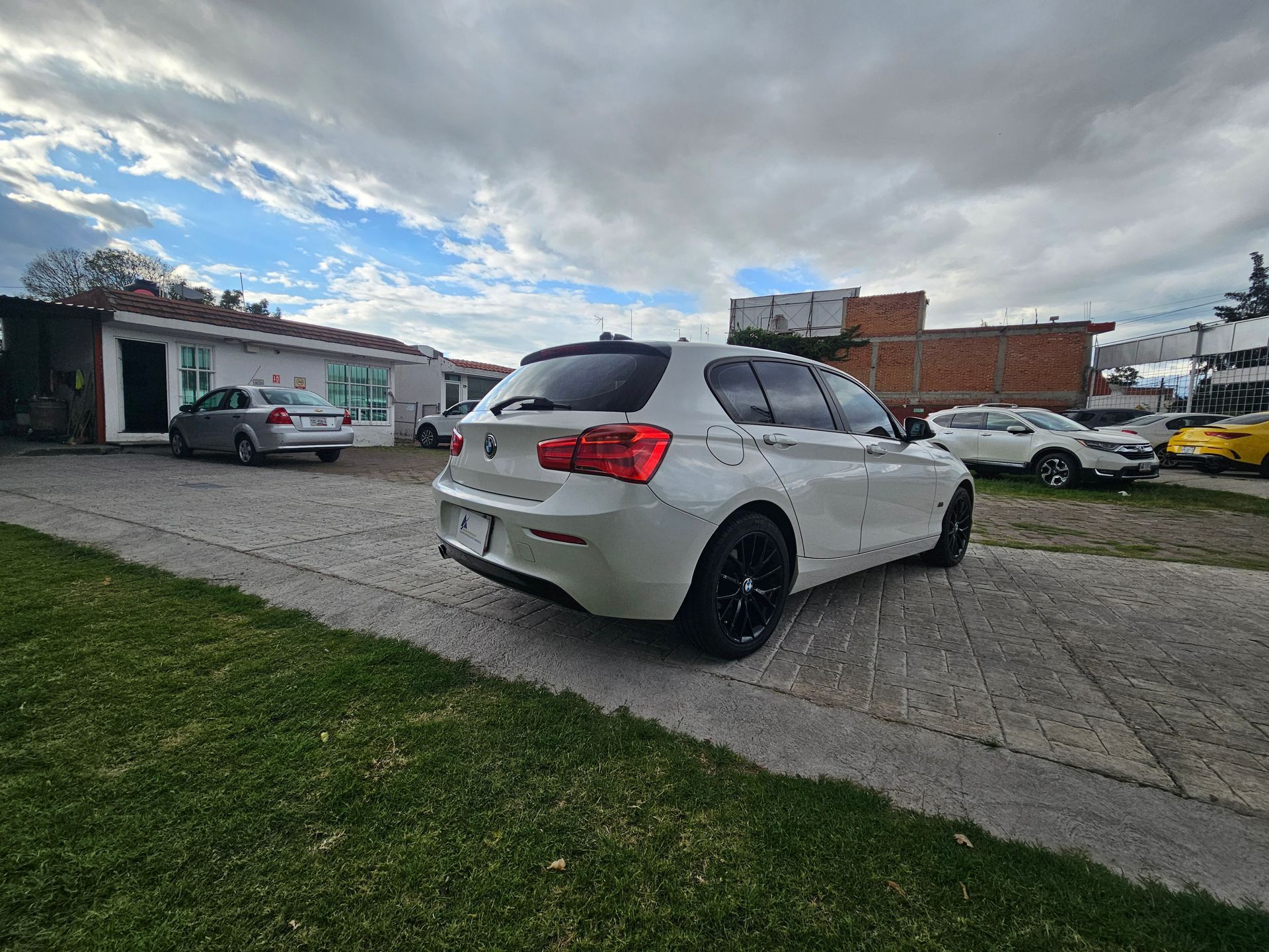 Un BMW hatchback blanco estacionado en un lote pavimentado con otros autos, cielo nublado.
