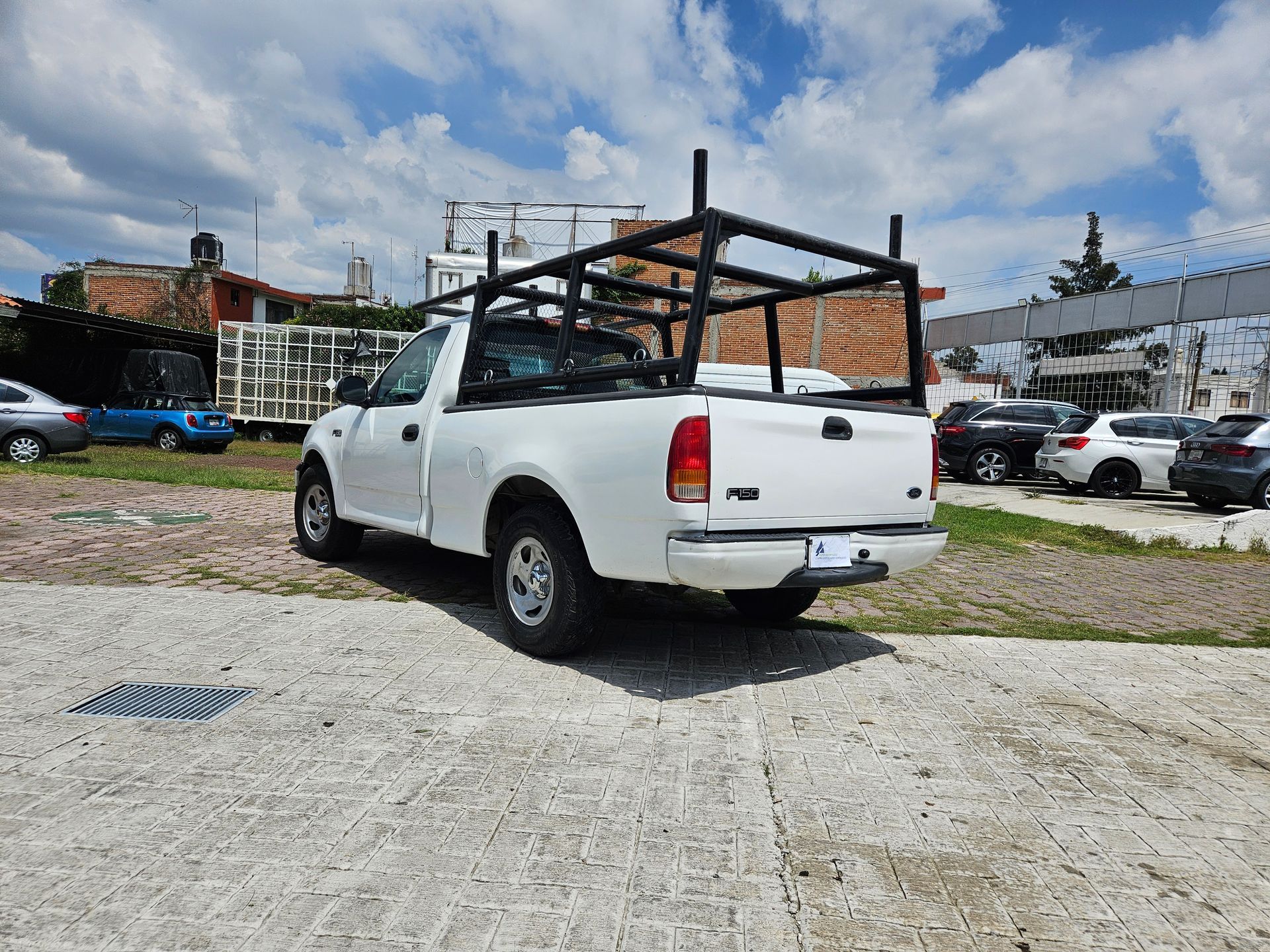 Camioneta blanca con portaequipaje negro estacionada sobre una superficie de concreto, varios autos al fondo.