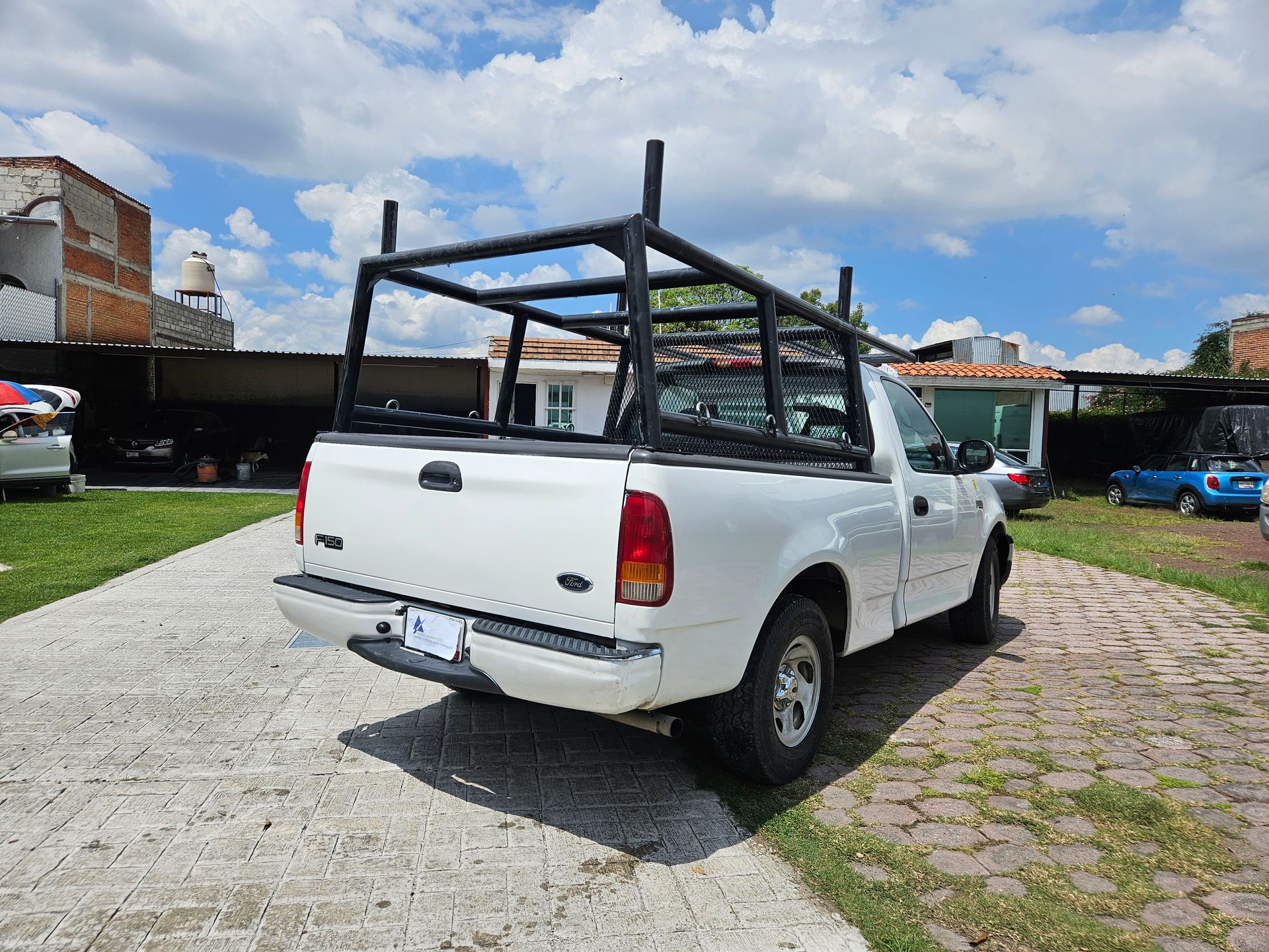 Camioneta blanca con parrilla negra estacionada en una entrada pavimentada, rodeada de edificios y vegetación.