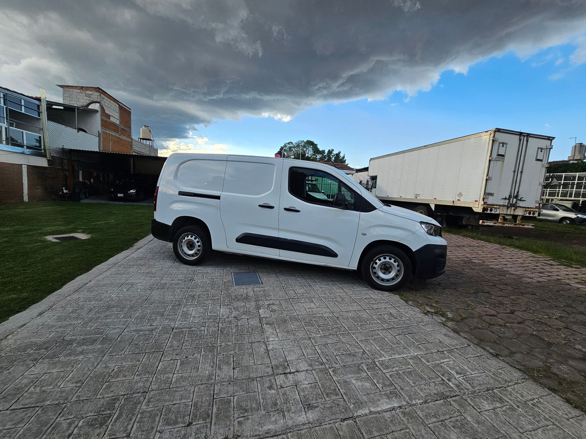 Furgoneta de carga blanca estacionada sobre una superficie de hormigón con un cielo nublado al fondo cerca de un remolque grande.