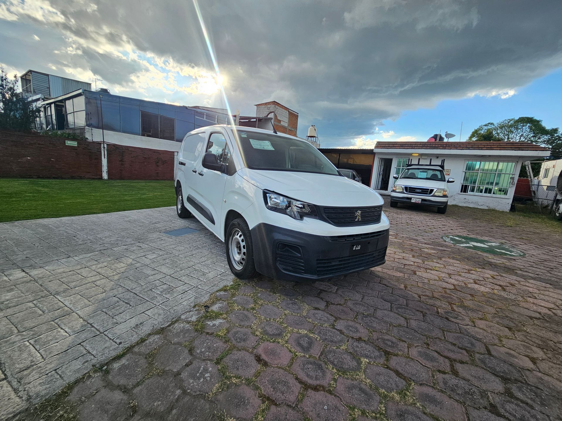 Furgoneta comercial blanca estacionada sobre adoquines con un pequeño edificio y otros vehículos al fondo. Cielo nublado.