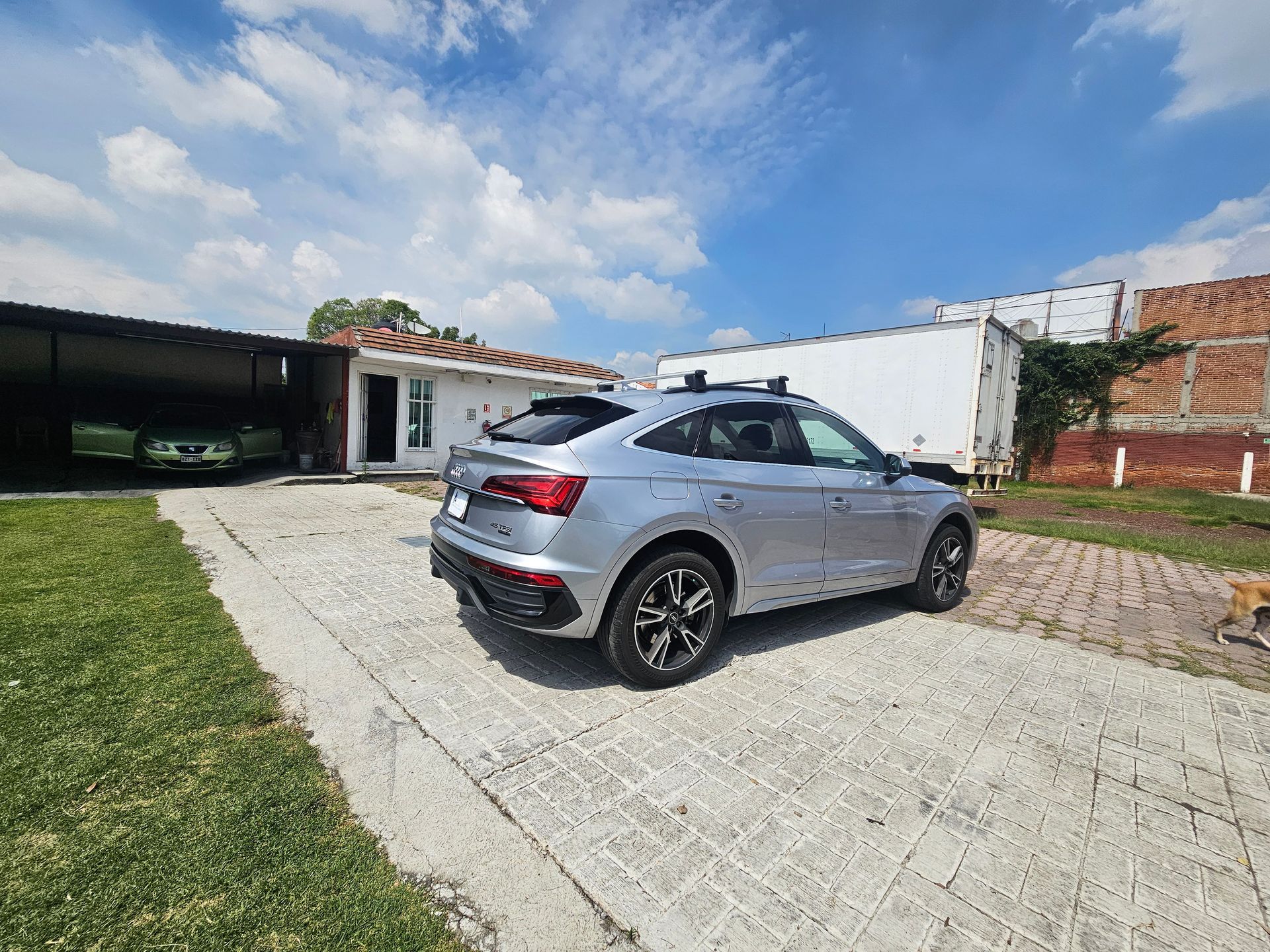 Una camioneta plateada estacionada en una entrada pavimentada frente a una casa en un día soleado. Un auto verde está en la cochera.