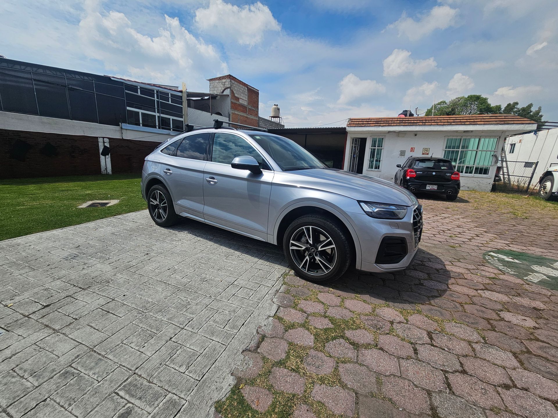 SUV Audi plateado estacionado en un camino pavimentado con un área de césped y un edificio al fondo en un día soleado.