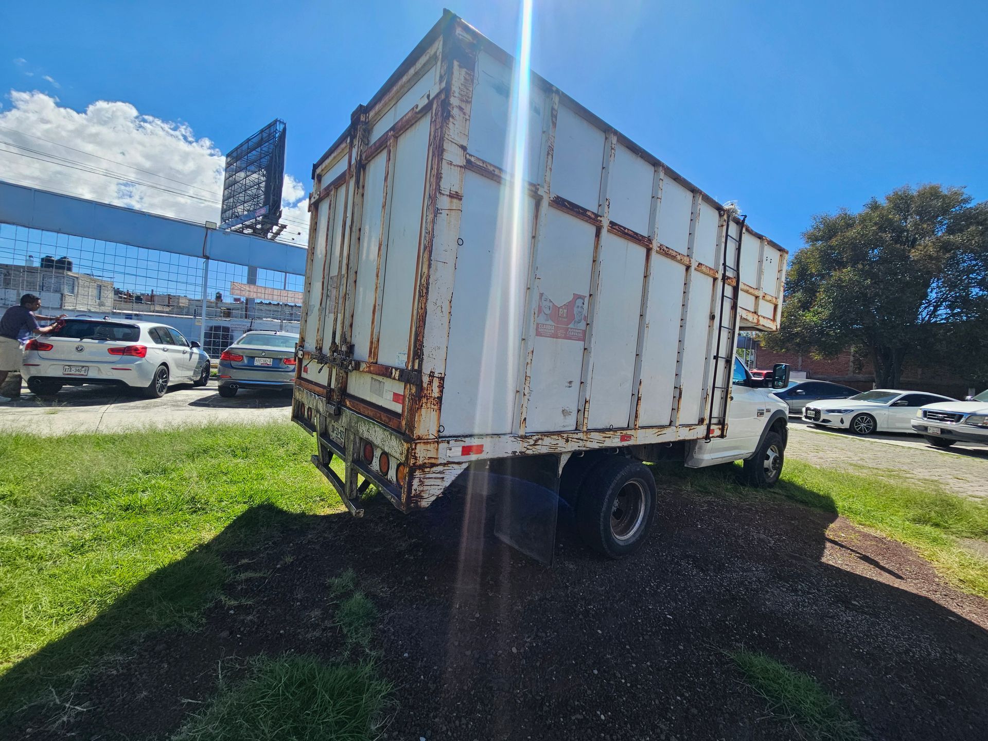 Camión blanco con caja de carga sobre césped, estacionado en un día soleado. Se ven otros vehículos y una valla publicitaria al fondo.