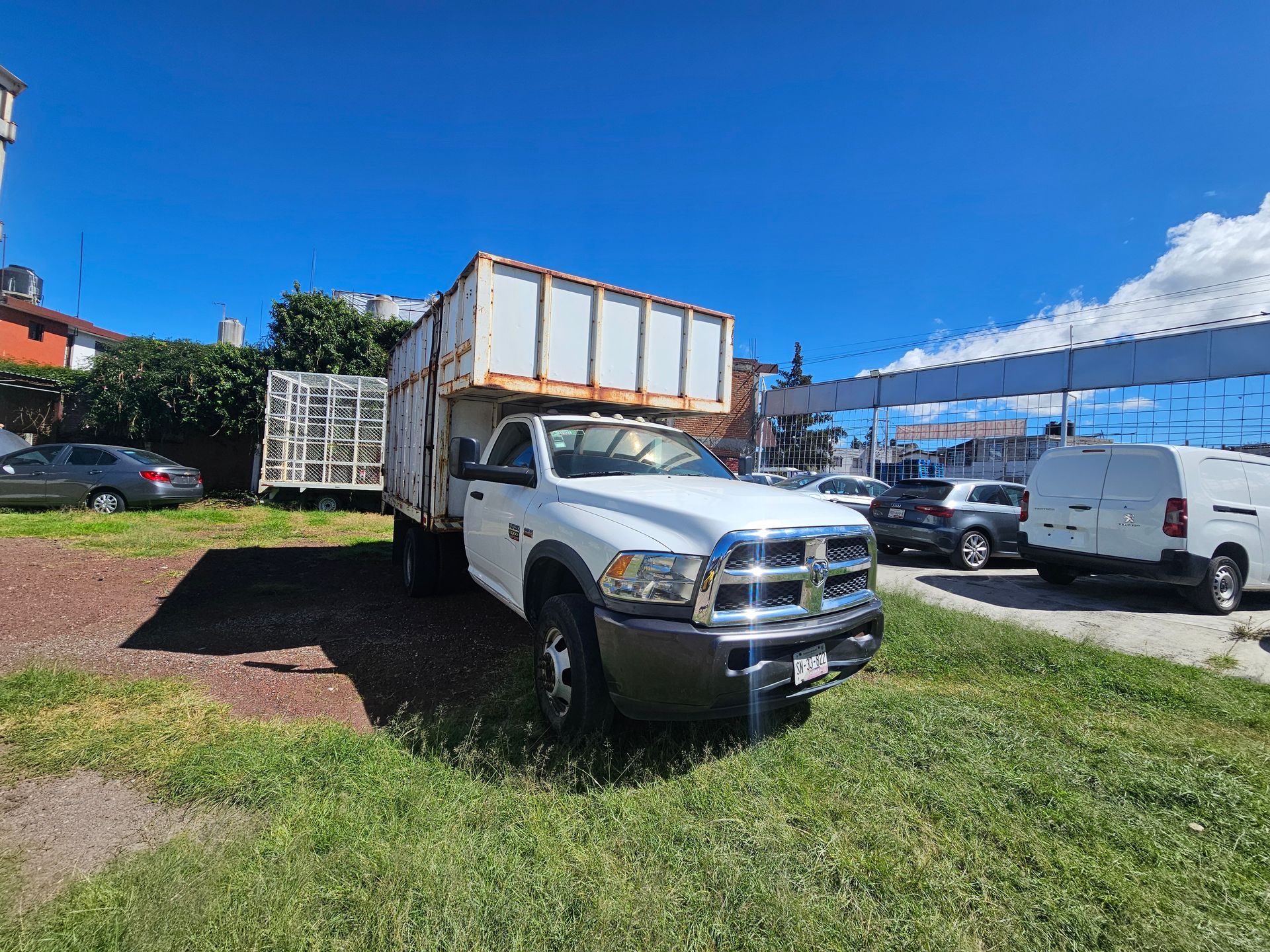 Camión Ram blanco con caja de carga de metal, estacionado en el césped bajo un cielo azul.