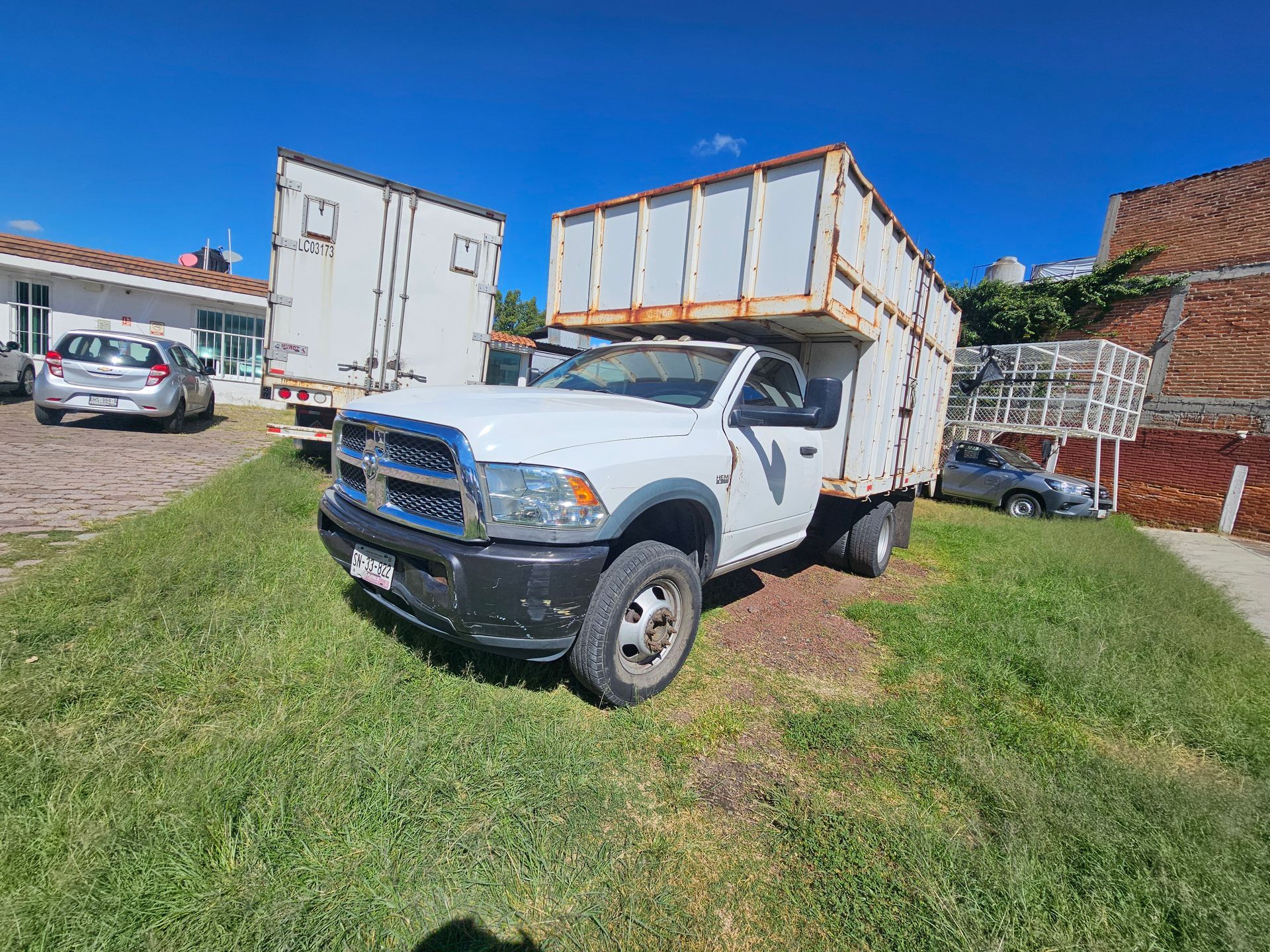 Camioneta Ram blanca con un amplio compartimento de carga, estacionada sobre césped. Otros vehículos y edificios al fondo.