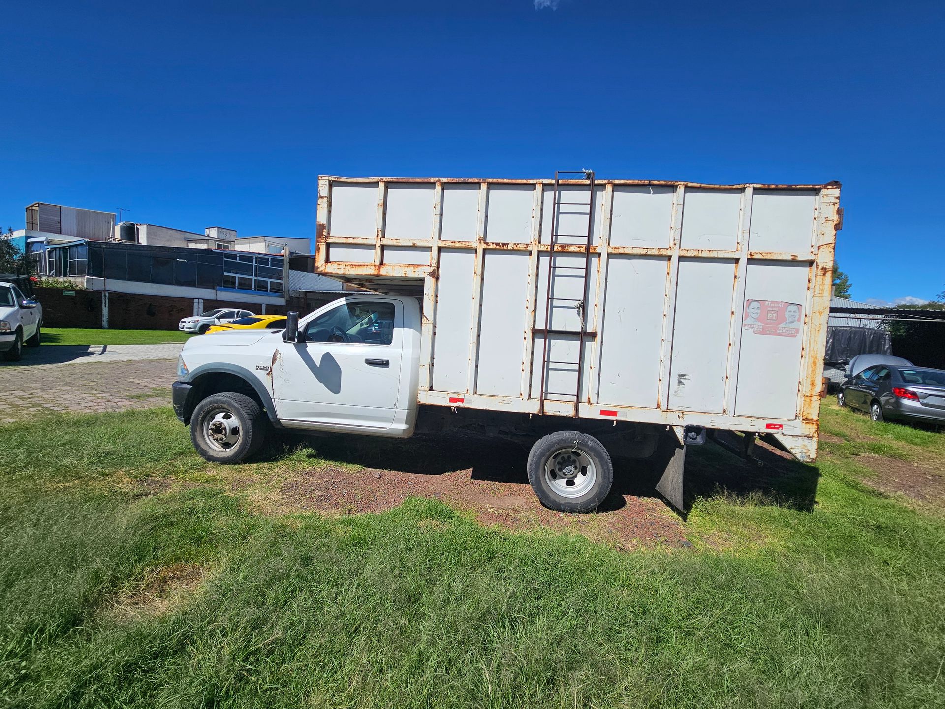 Camión blanco con caja de carga alta estacionado en el césped bajo un cielo azul claro.