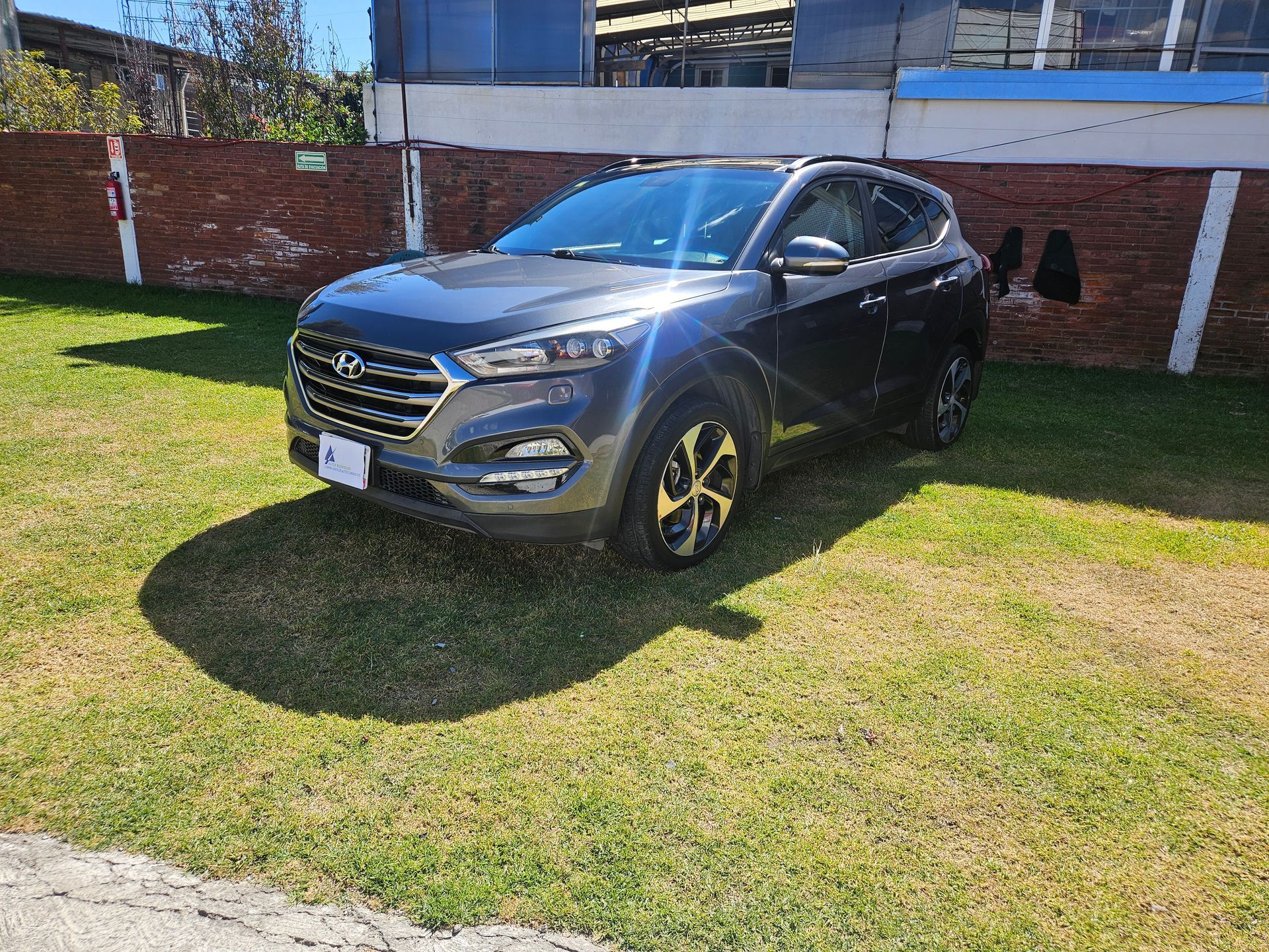 Hyundai Tucson gris oscuro estacionado en el césped, frente a una pared de ladrillos y edificios industriales, en un día soleado.