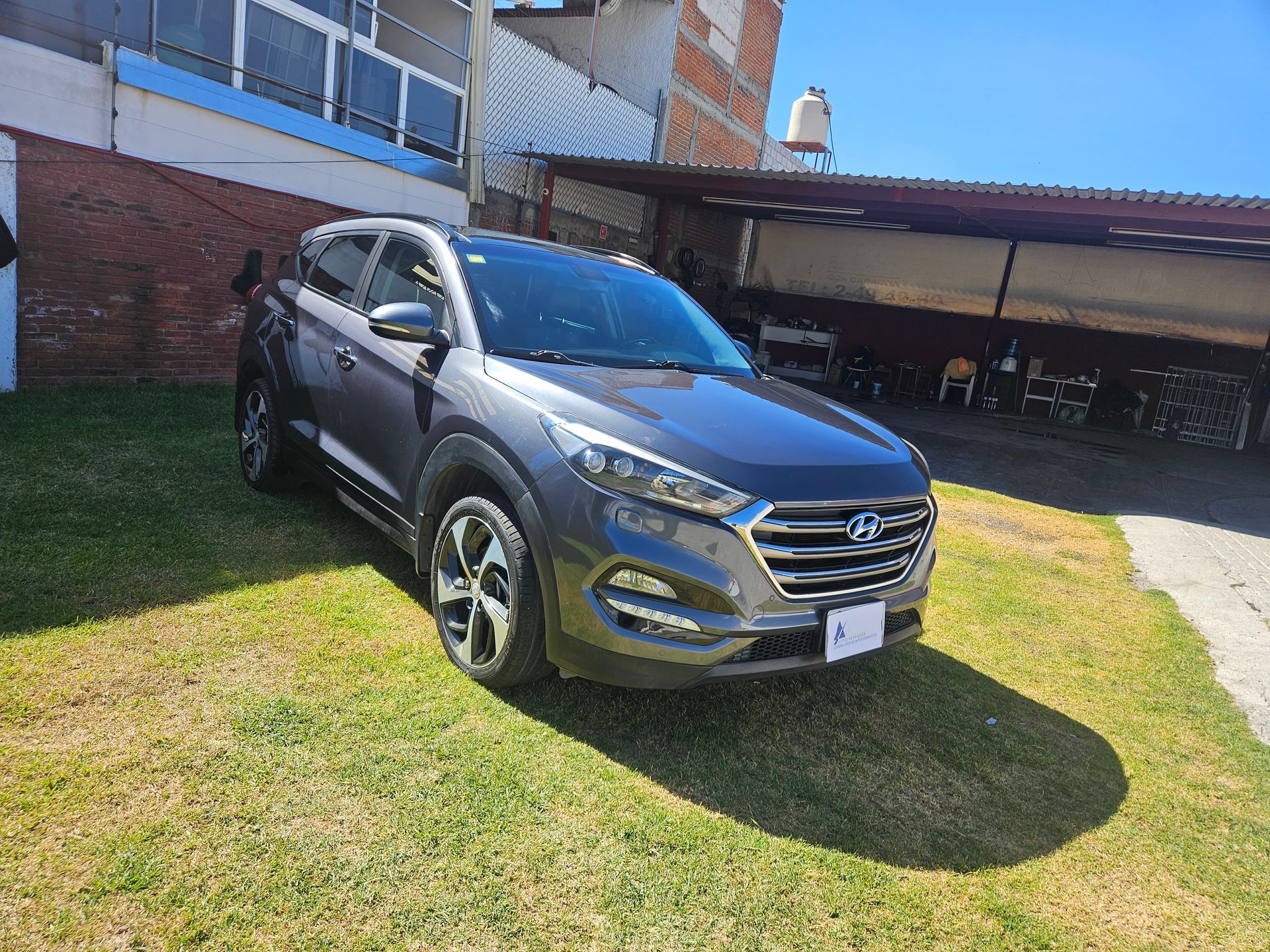 SUV Hyundai Tucson gris estacionado en el césped frente a un edificio de ladrillos.