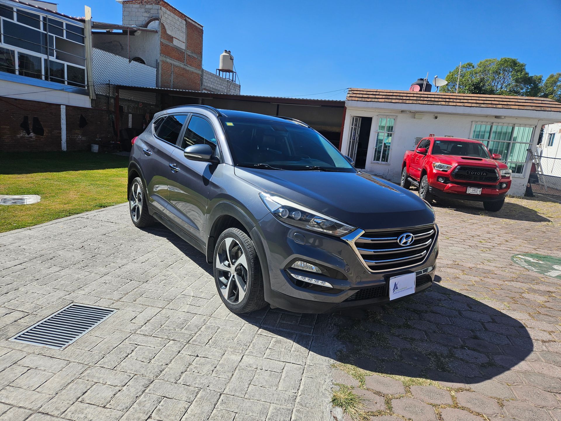 SUV Hyundai Tucson gris estacionado en una entrada pavimentada; camioneta roja al fondo.