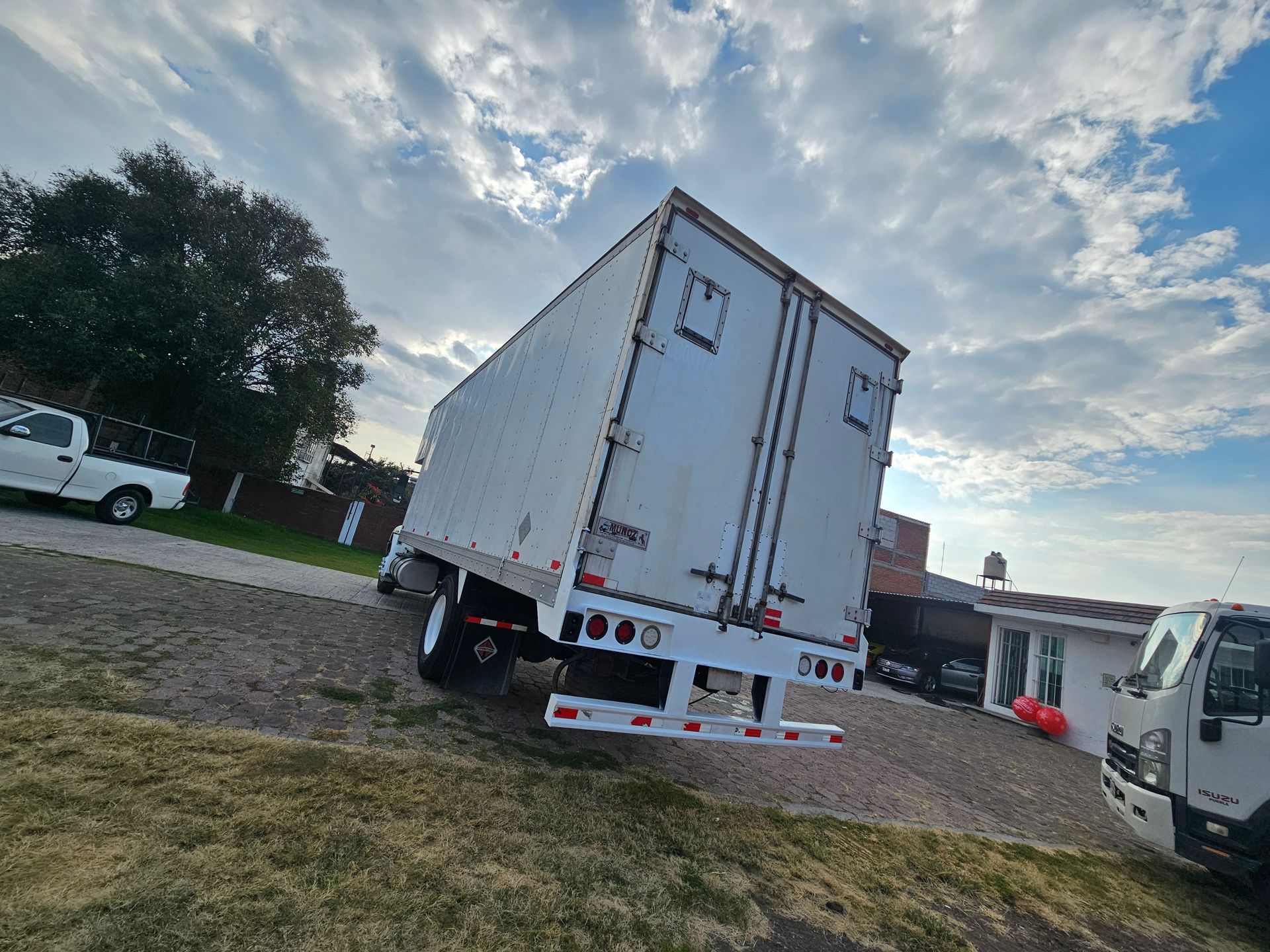 Camión de caja blanca estacionado sobre una superficie de grava al lado de una zona cubierta de hierba con un cielo nublado de fondo.