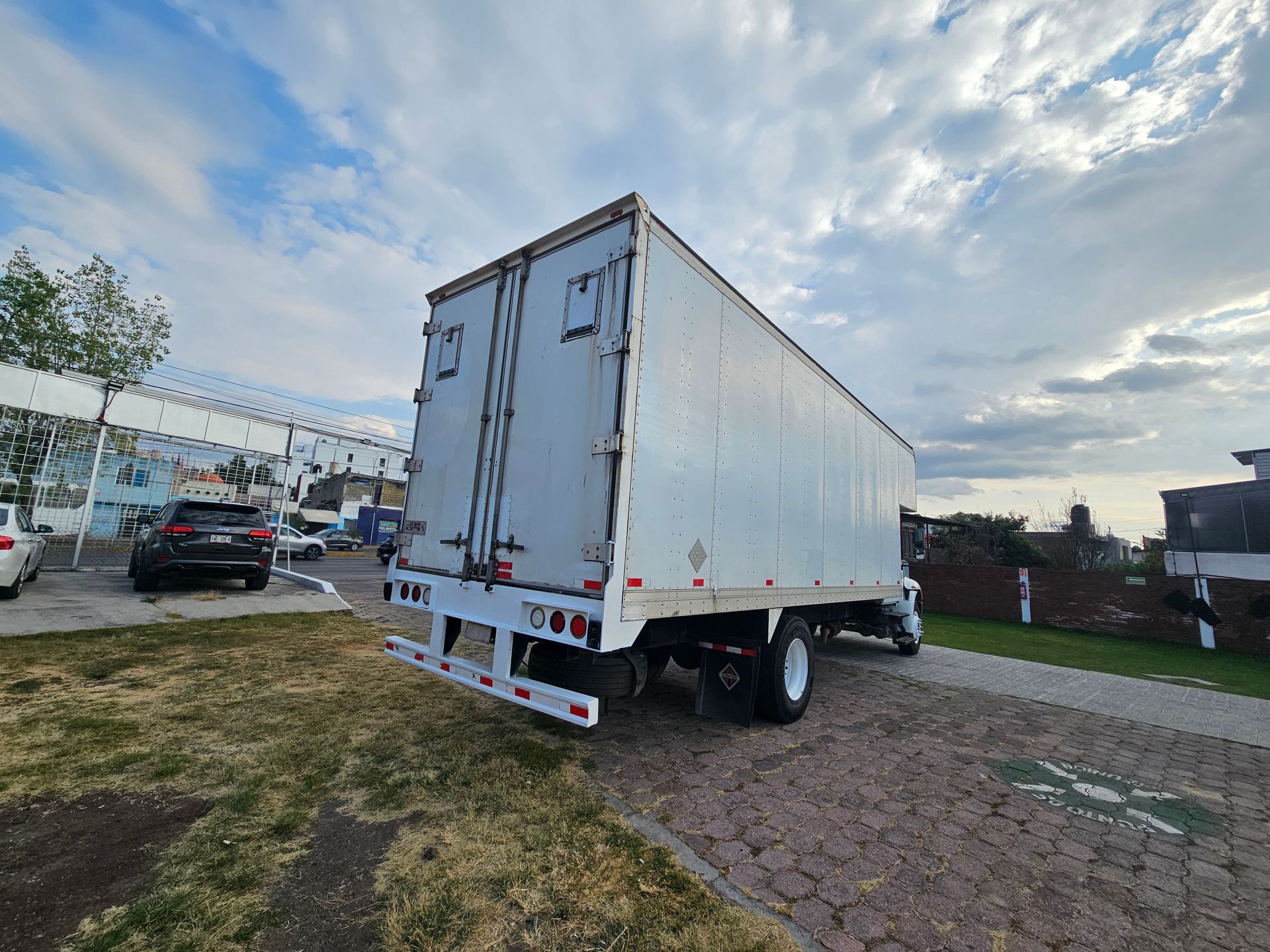 Vista trasera de un camión semirremolque blanco estacionado sobre pavimento y césped bajo un cielo nublado.