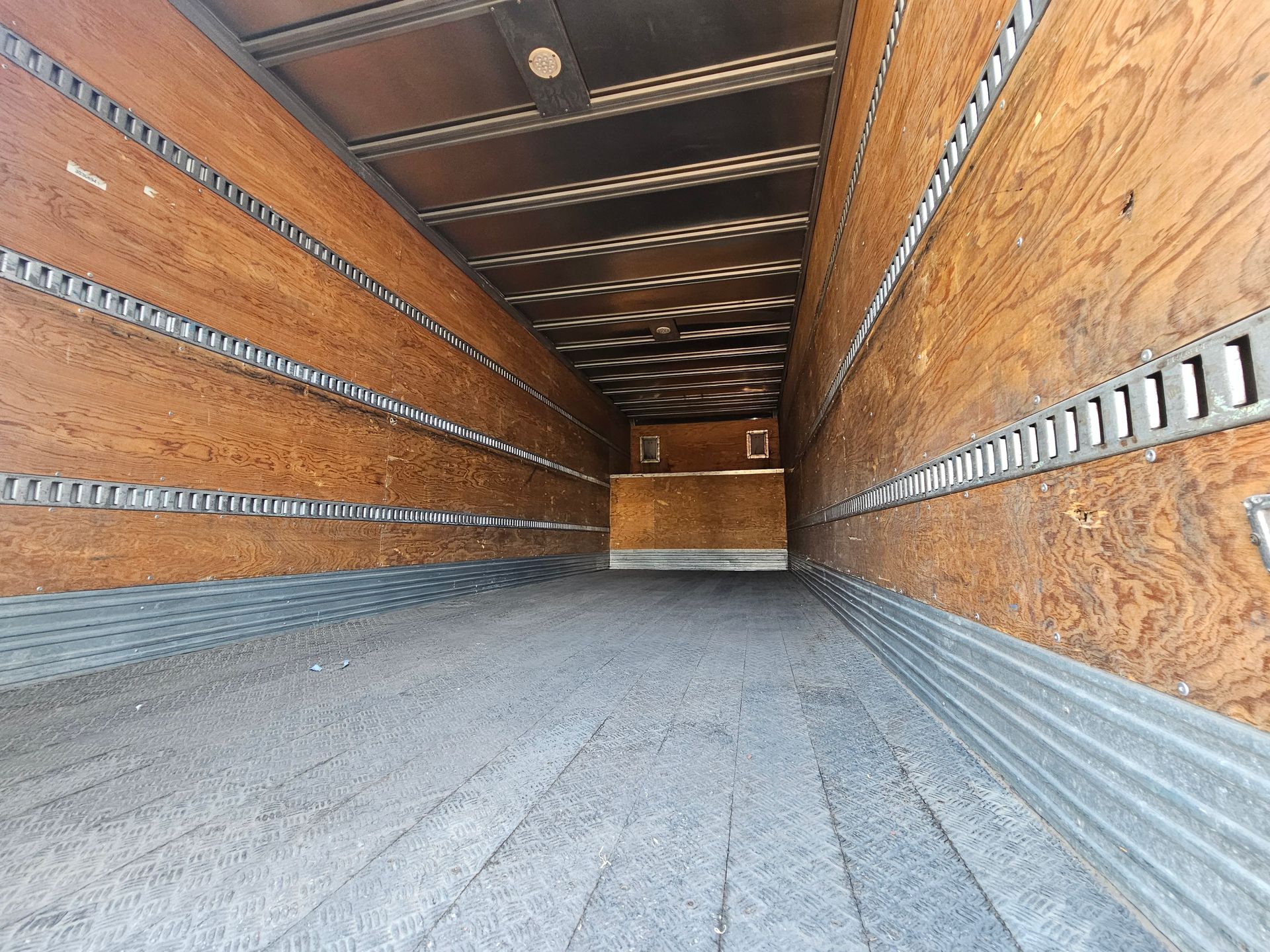 Interior de un semirremolque vacío. Paredes revestidas de madera con rieles de amarre de carga, suelo y techo grises.