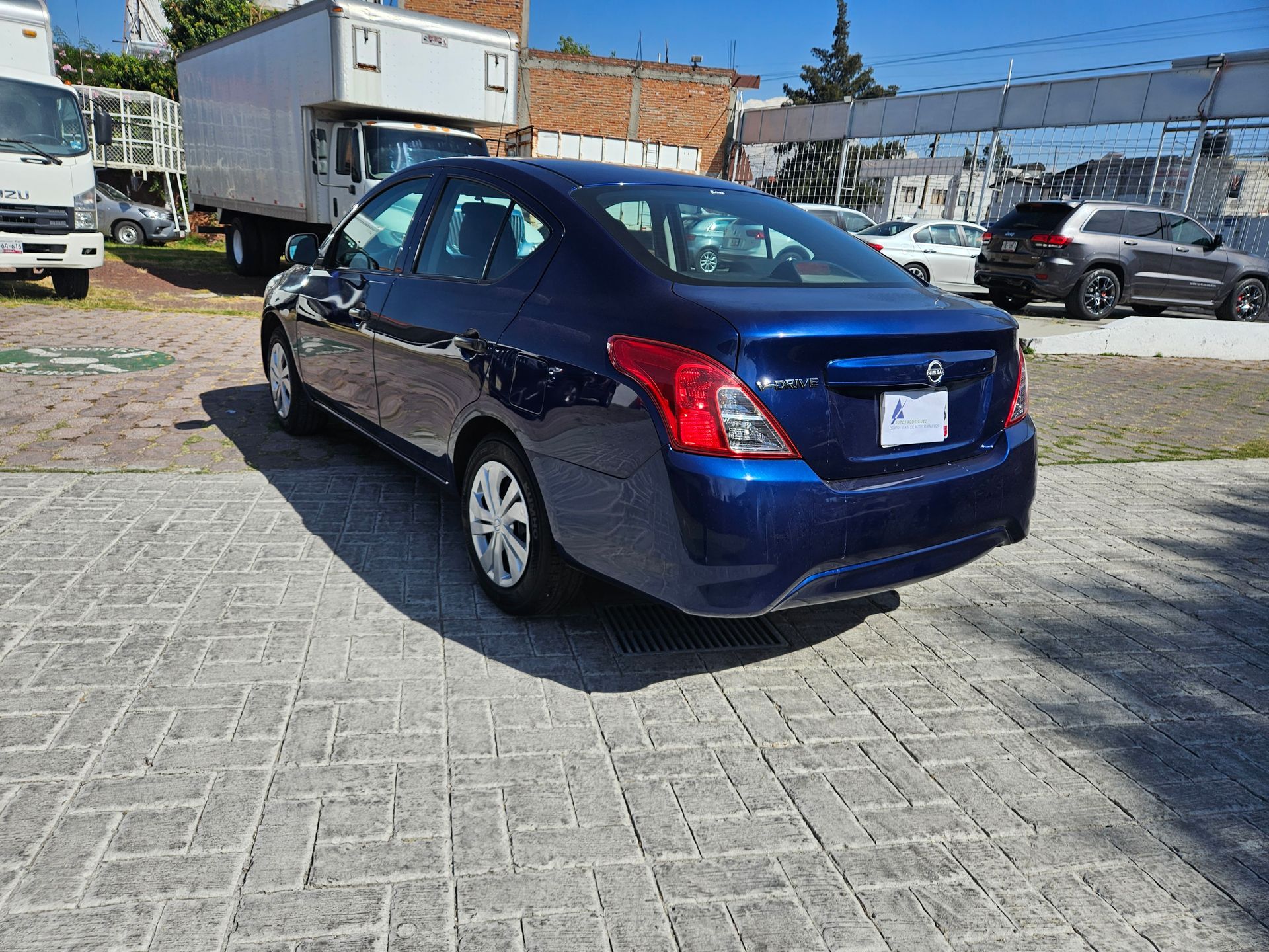 Sedán azul estacionado sobre adoquines de ladrillo, con camiones y otros vehículos visibles al fondo.