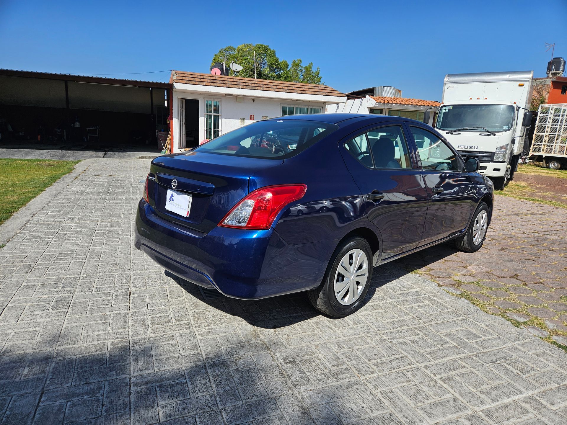 Un sedán Nissan azul estacionado en una entrada pavimentada frente a un edificio y un camión blancos.
