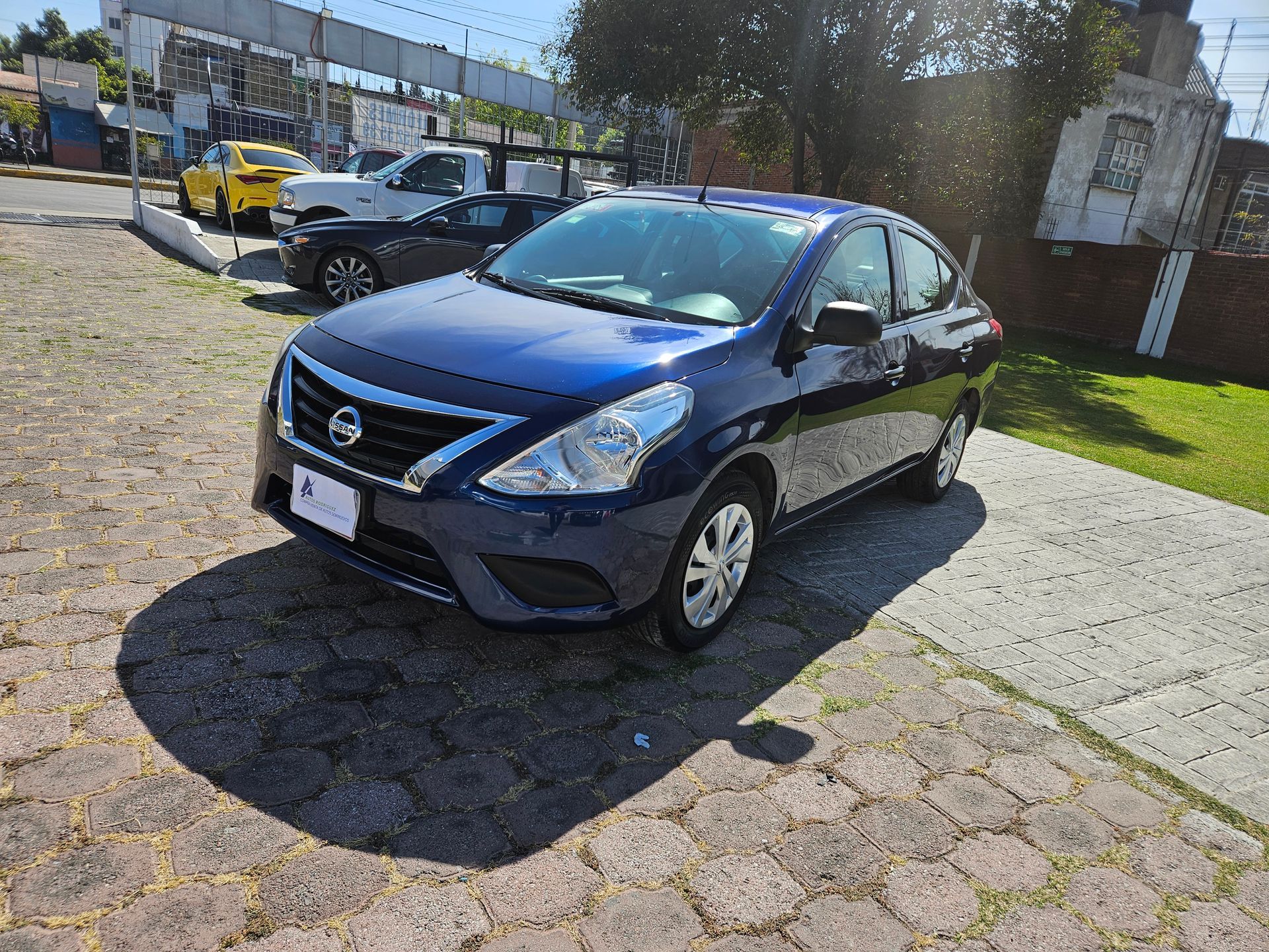 Un Nissan Versa sedán azul está estacionado sobre una superficie de ladrillo. La luz del sol proyecta sombras. Hay otros vehículos al fondo.