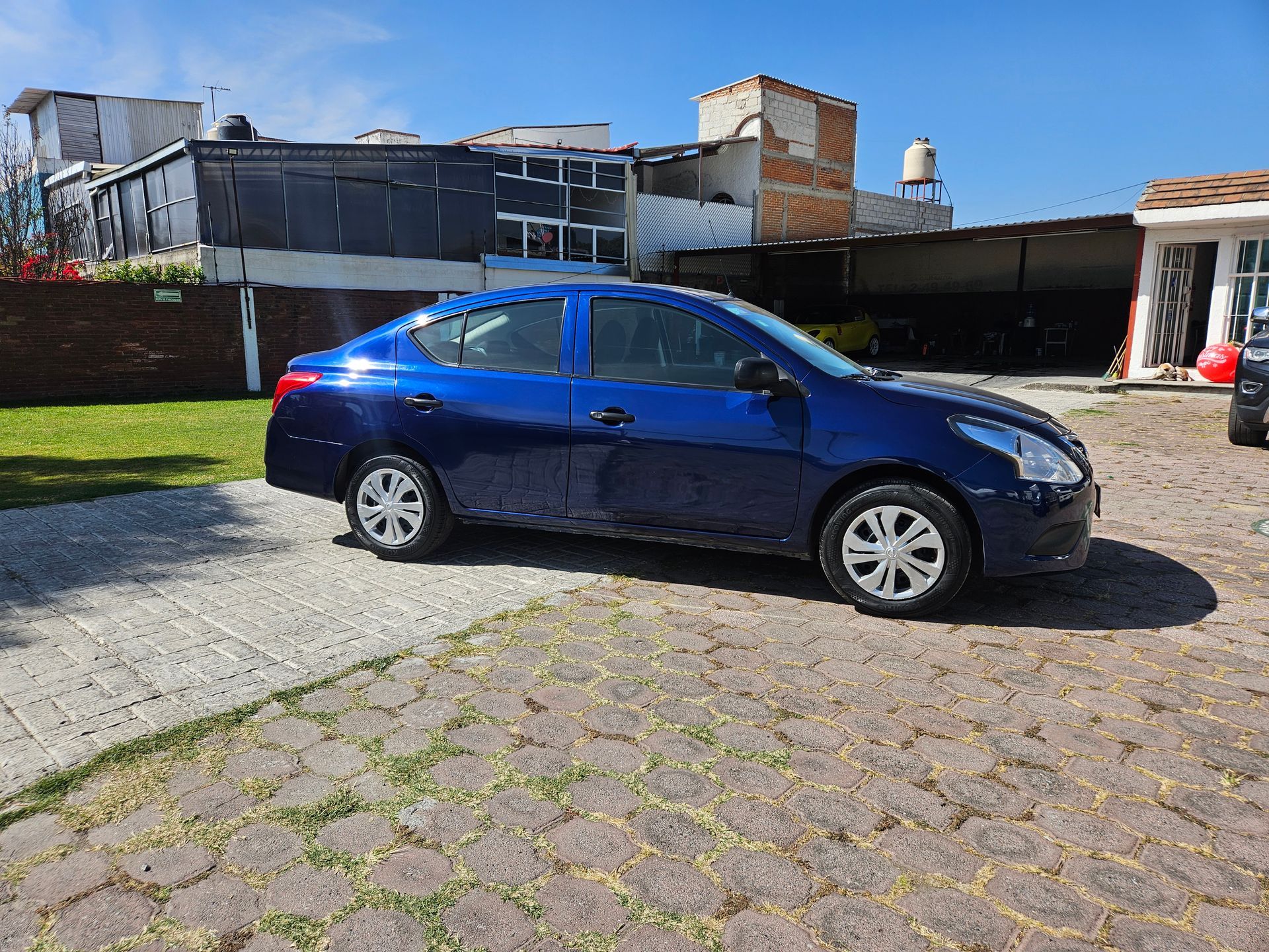 Sedán azul estacionado en un camino de piedra, con edificios y un patio cubierto de césped al fondo.