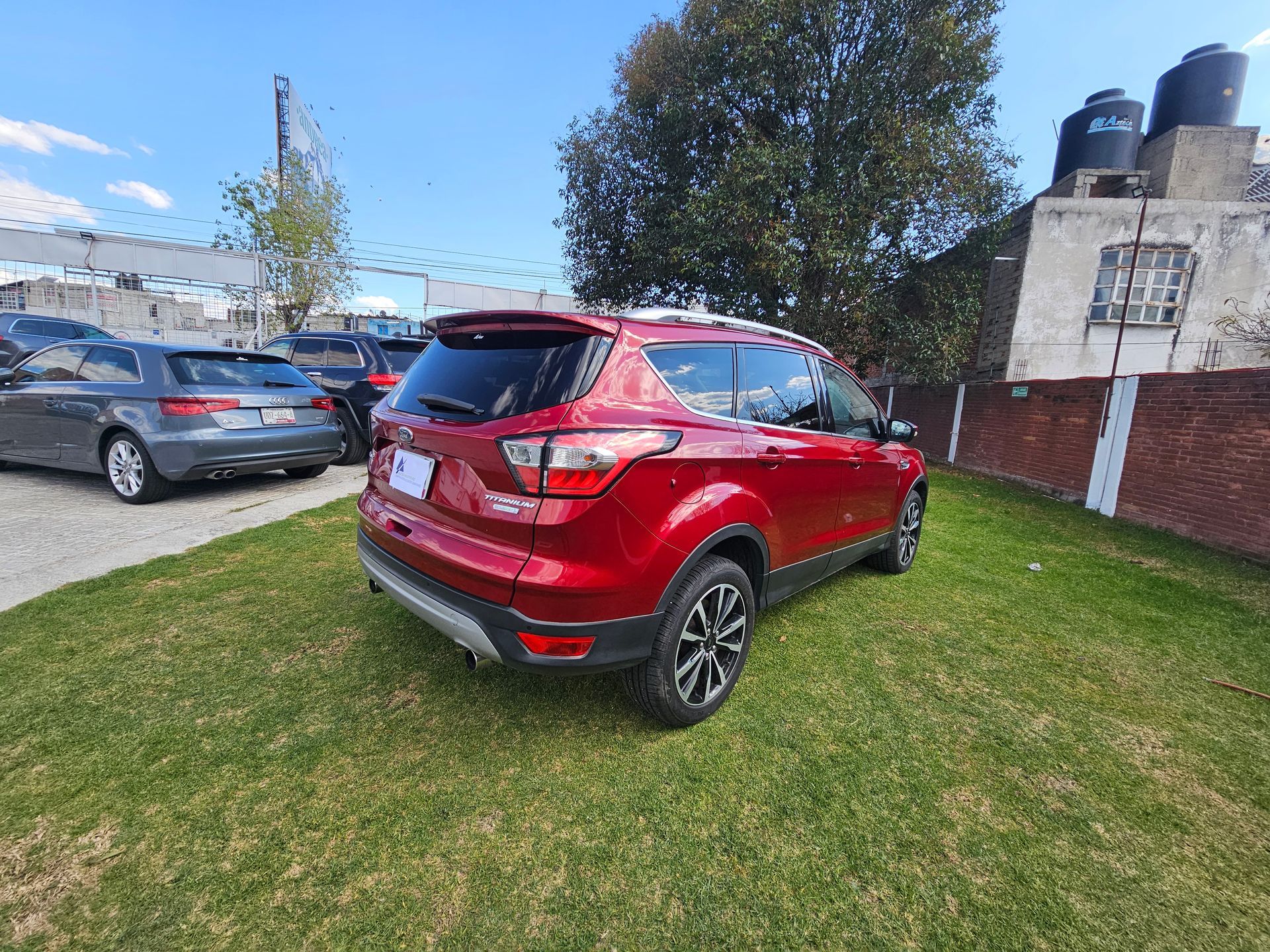Una camioneta Ford Escape roja estacionada sobre césped verde. Otros autos y edificios al fondo.