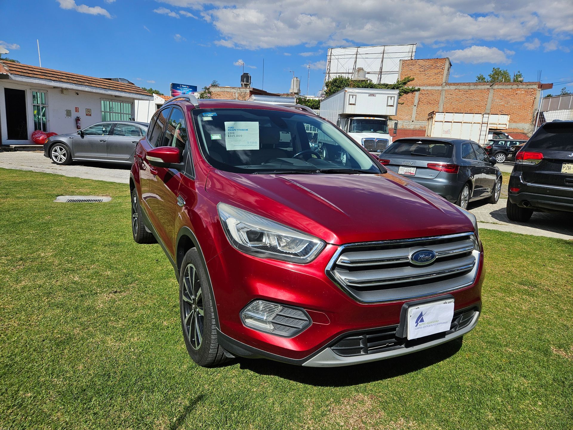 SUV Ford Escape rojo estacionado sobre el césped, con otros vehículos y edificios al fondo en un día soleado.