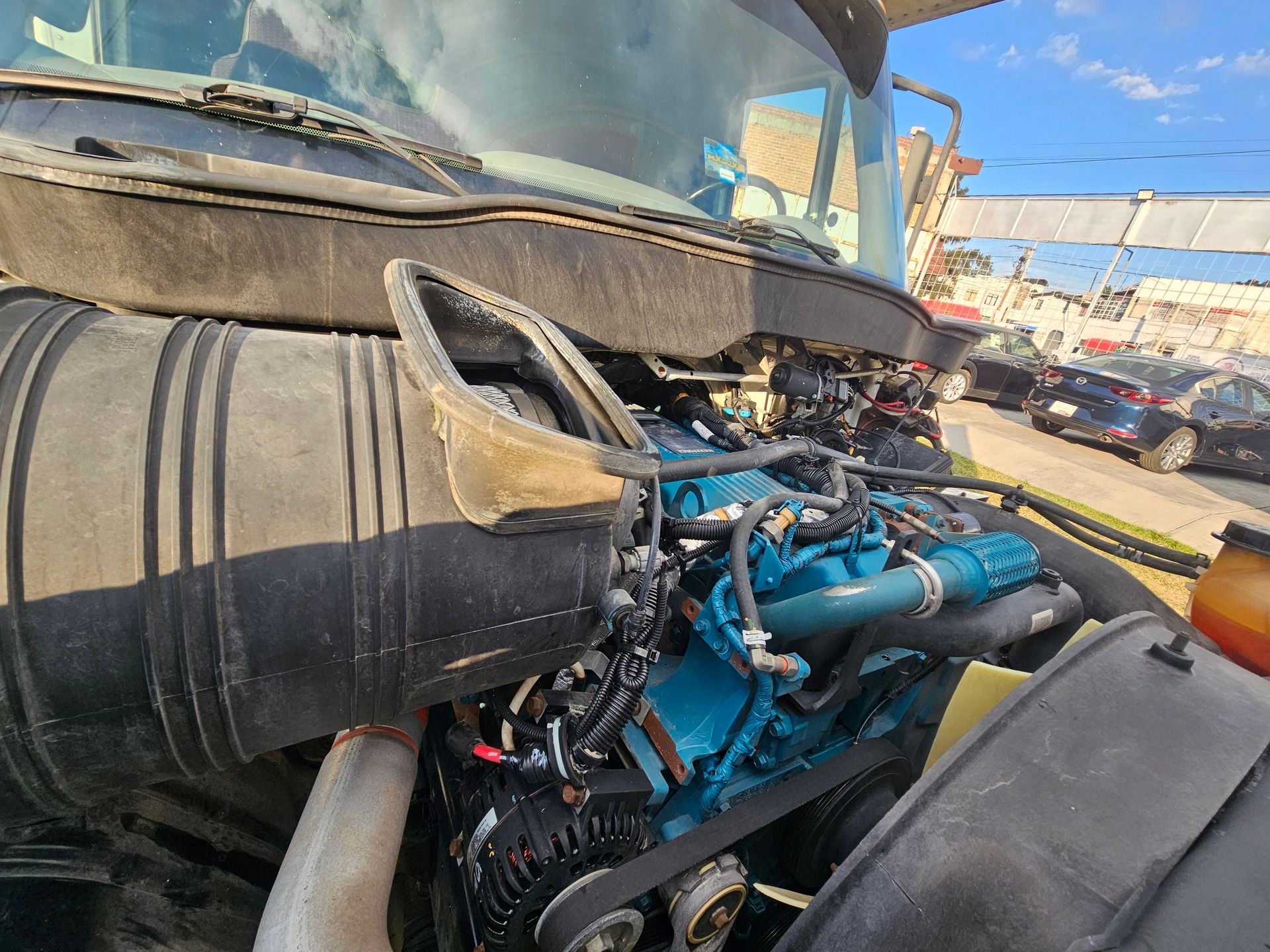 Capó abierto de un camión semirremolque que muestra un motor azul sobre un fondo de vehículos y un estadio.