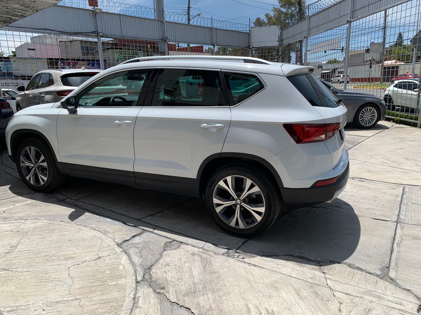 Vehículo todo terreno de color blanco estacionado sobre una superficie de concreto, frente a otros vehículos y edificios.