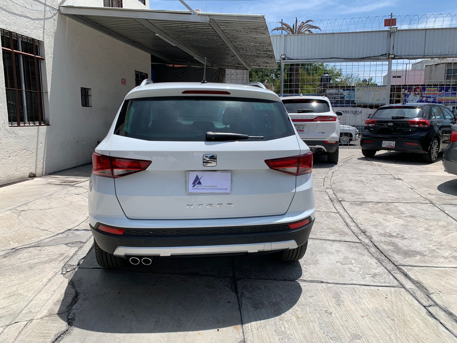 SUV blanca estacionada al aire libre, vista trasera. Se ven otros vehículos y el edificio. Día soleado.