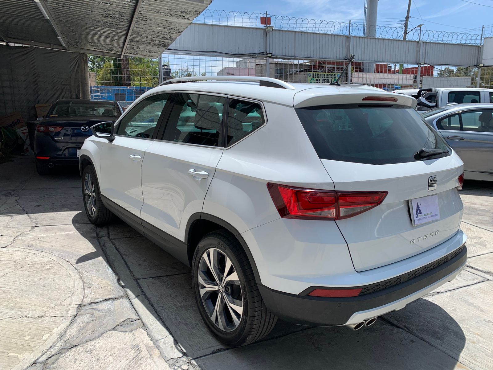 SUV blanco estacionado bajo una marquesina. Se muestra la vista trasera del vehículo, con luces, ruedas y ventanas visibles.