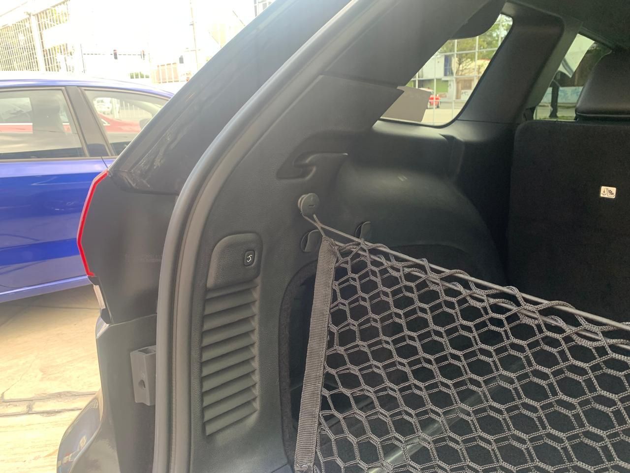 Interior de baúl de automóvil negro con red de carga y ventilaciones; automóvil azul parcialmente visible.