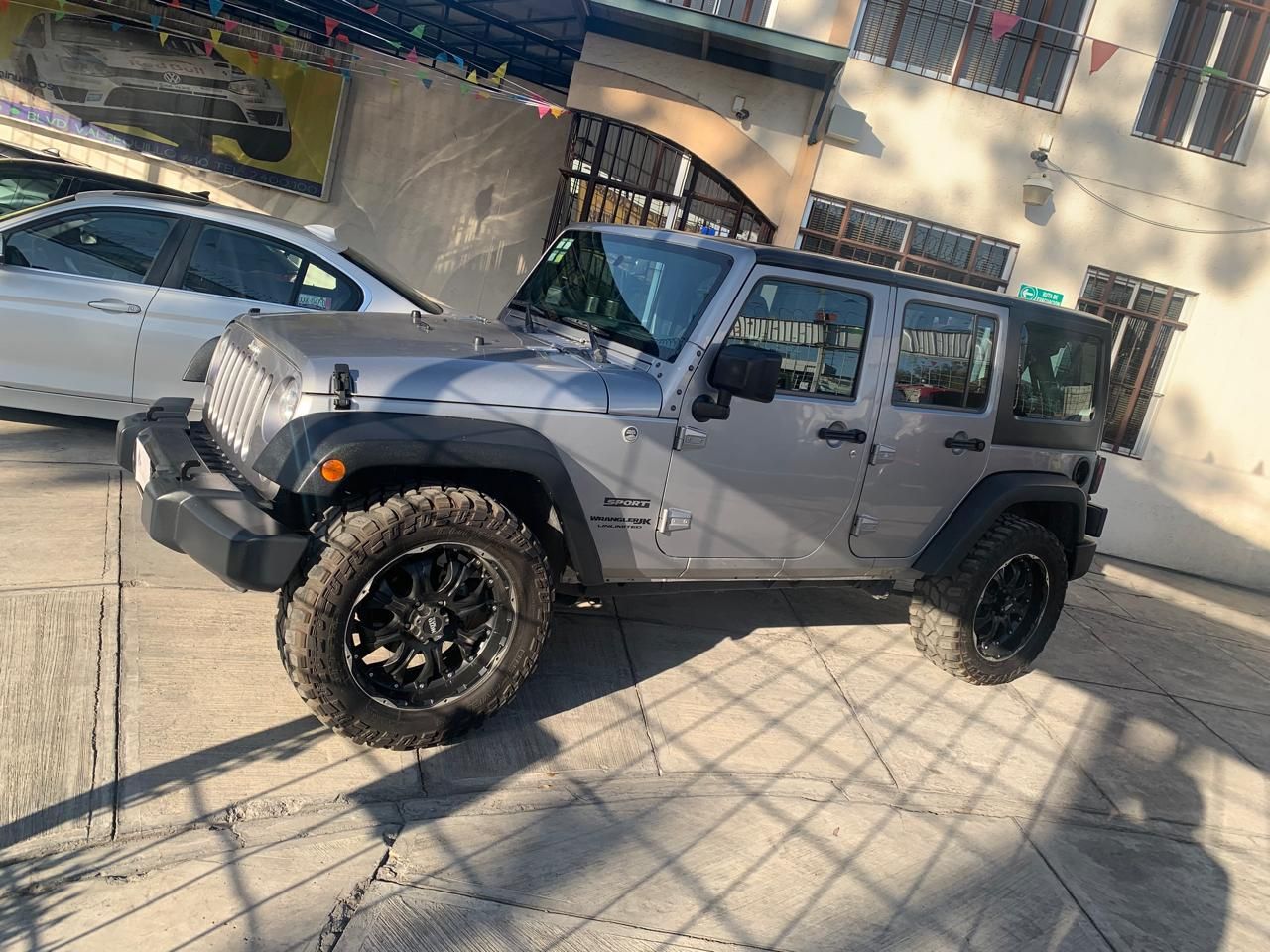 Jeep Wrangler plateado estacionado frente a un edificio; ruedas y neumáticos negros.