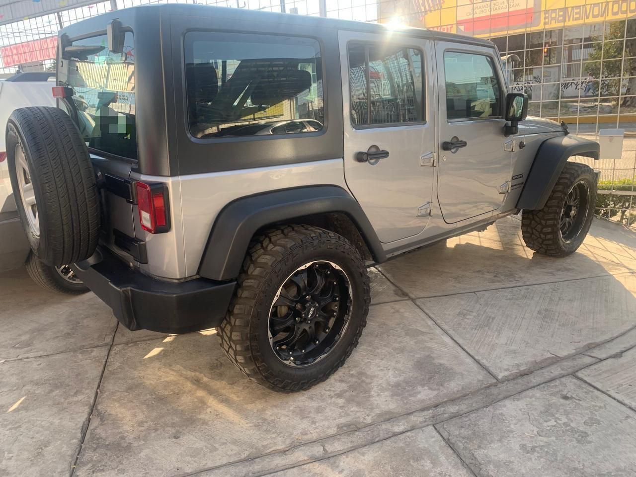 Jeep Wrangler plateado estacionado sobre concreto. Rines negros, llanta de repuesto, techo negro, día soleado.