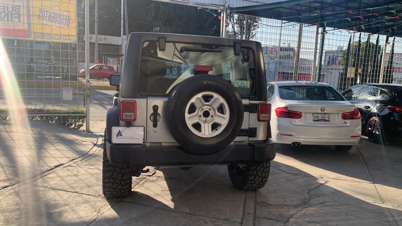 Jeep Wrangler plateado estacionado, vista trasera, llanta de repuesto, techo negro y autos estacionados al fondo.