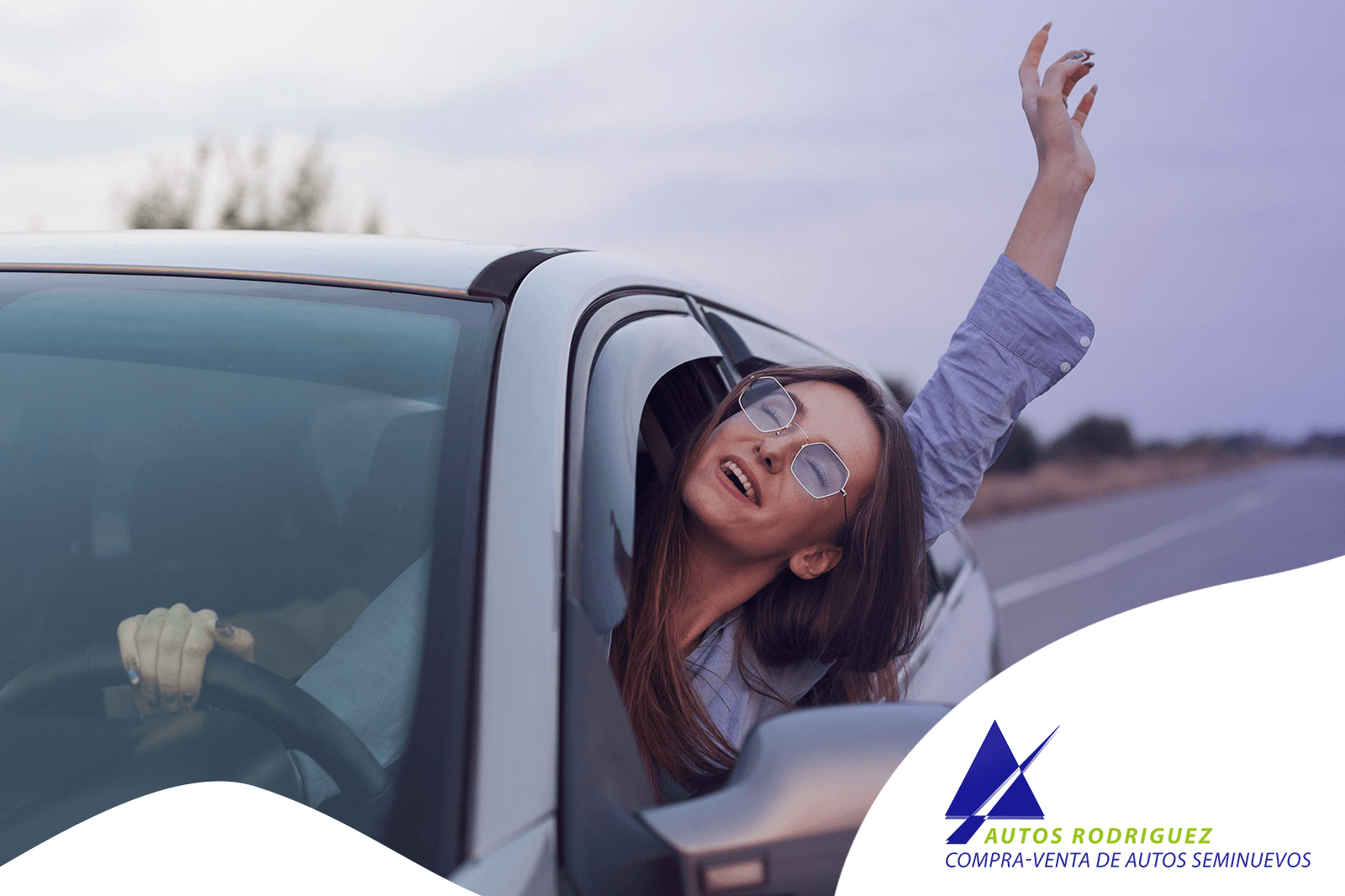 Mujer con gafas asomada a la ventanilla de un coche, con el brazo levantado y sonriendo.