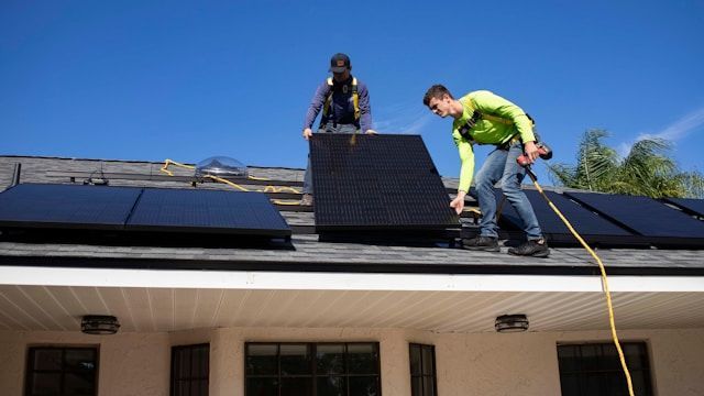 Two workers installing black solar panels on a roof under a clear blue sky