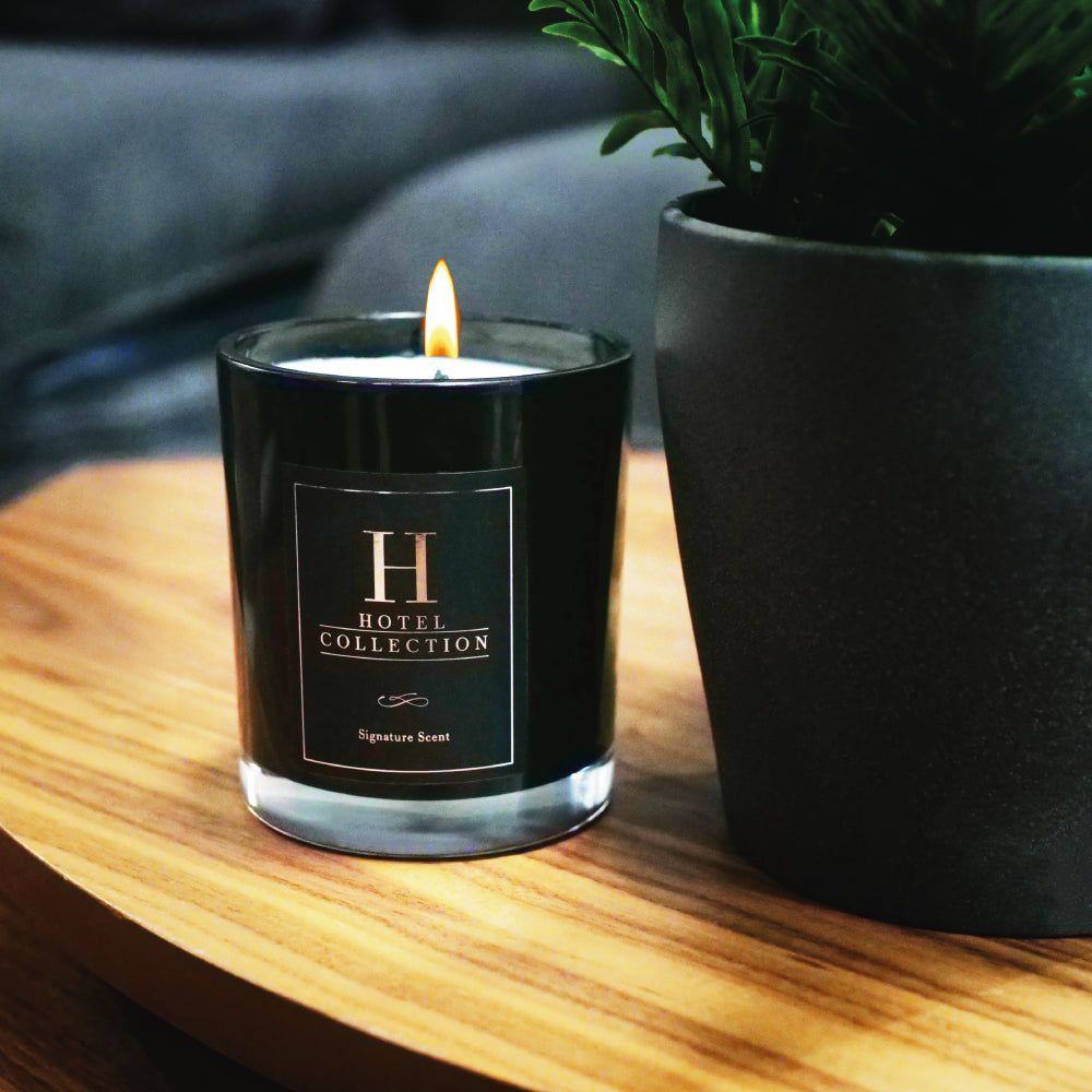 A hotel collection candle is sitting on a wooden table next to a potted plant.