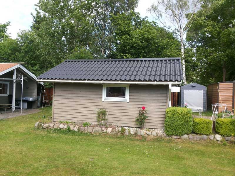 Lille gråt skur med mørkt tag og vindue i en grøn gård med en rosenbusk.