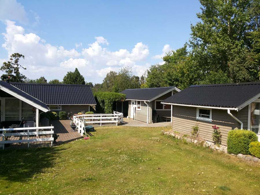 Adskillige grå bungalows med sorte tage på en græsklædt plæne under en blå himmel med træer.