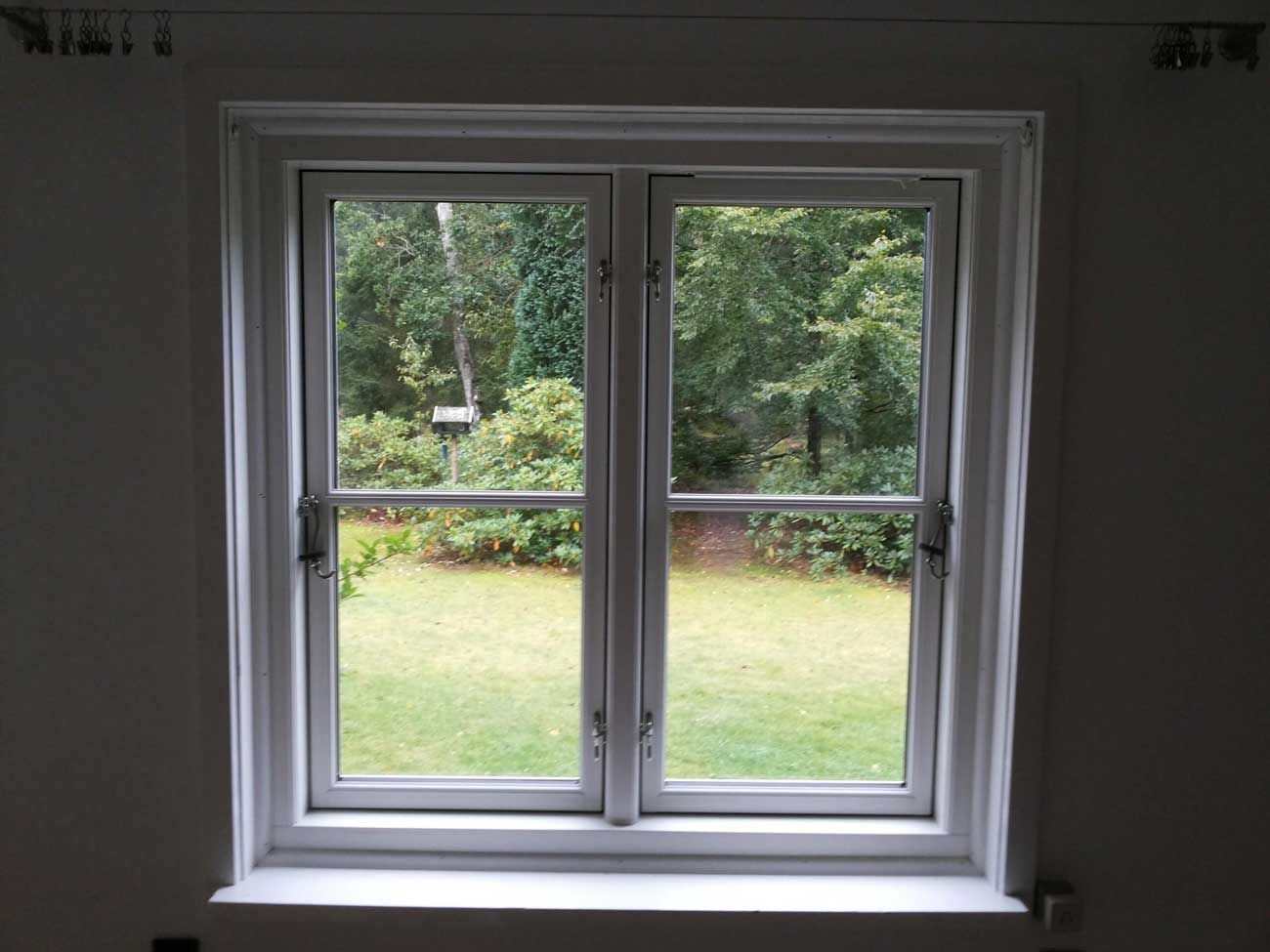 Hvidindrammet dobbeltvindue med udsigt til en grøn gårdhave med træer og buske.