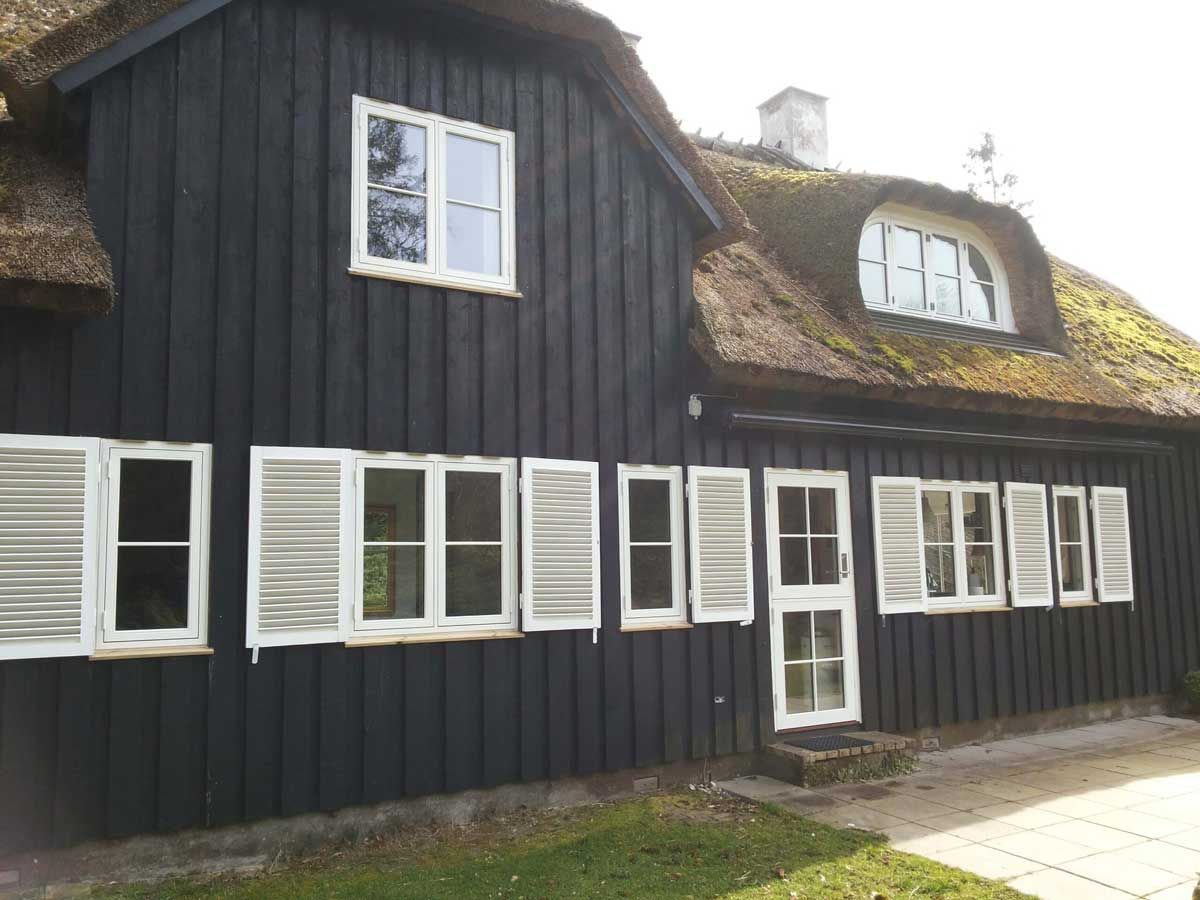 Sort træhus med hvide vinduesrammer og skodder, toppet med stråtag.