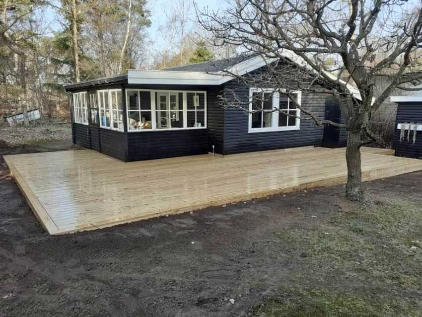 Sort hytte med træterrasse i en gård, træ til højre, overskyet dag.