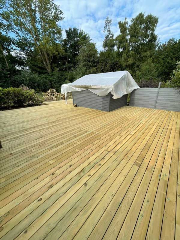 Træterrasse med gråt skur og træer i baggrunden under en overskyet himmel.