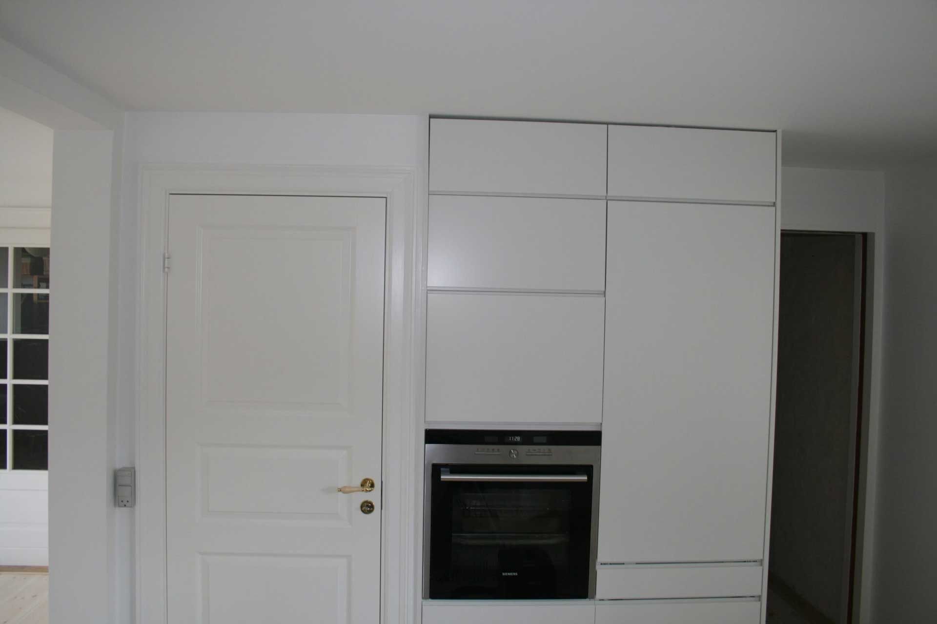 Hvide køkkenskabe med indbygget ovn og låge til venstre og en døråbning til højre.