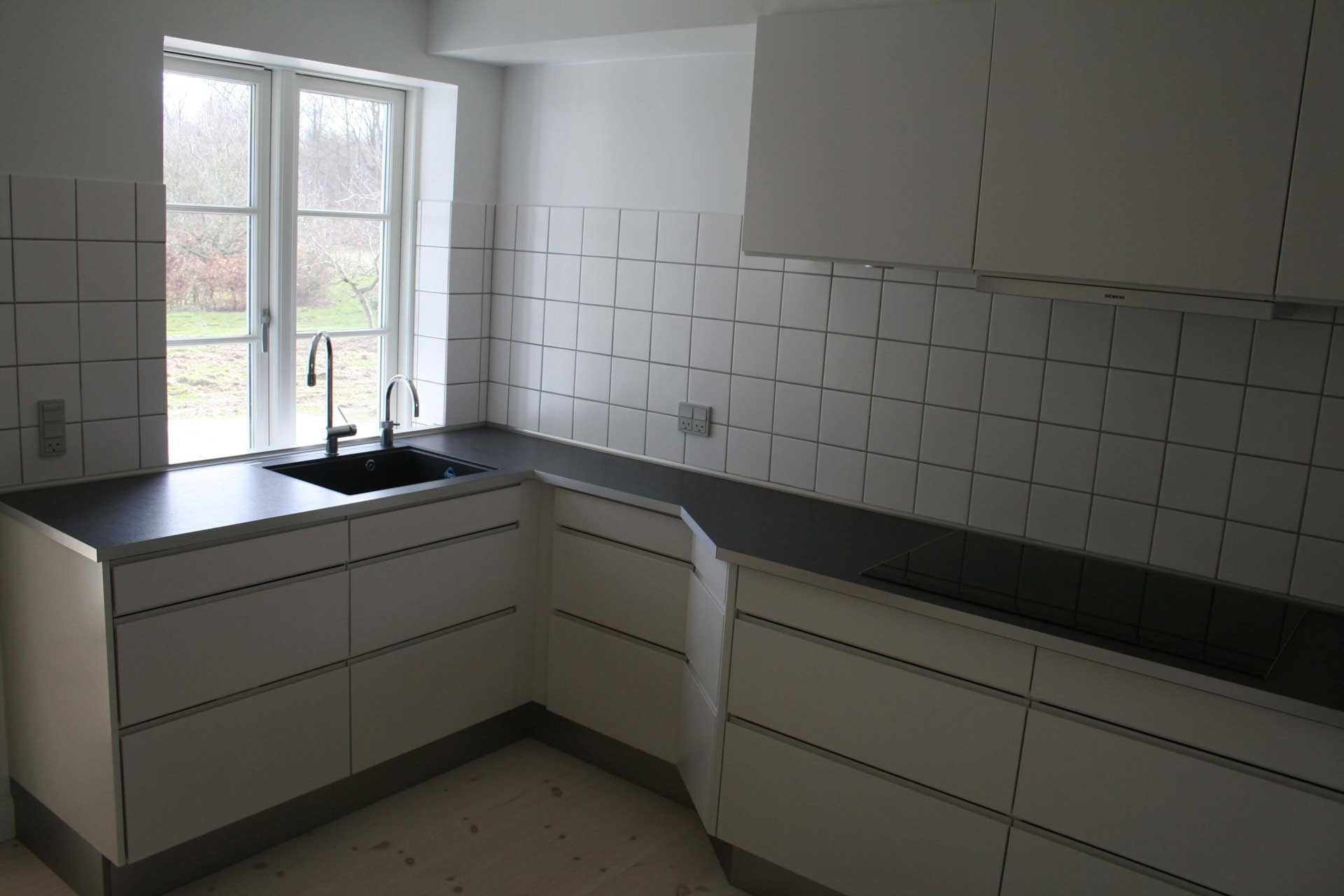 Køkken med hvide skabe, mørke bordplader og flisebelagt bagplade samt vindue og vask.