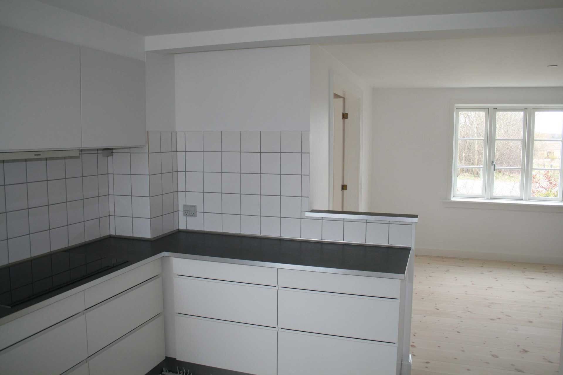Hvidt køkken med sorte bordplader, flisebelagt bagplade og et vindue i det tilstødende rum.