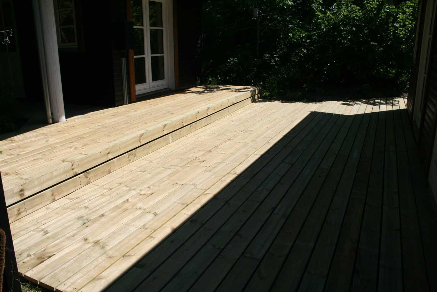 Træterrasse med trapper ved siden af ​​en bygning og træer. Sollys kaster lange skygger.