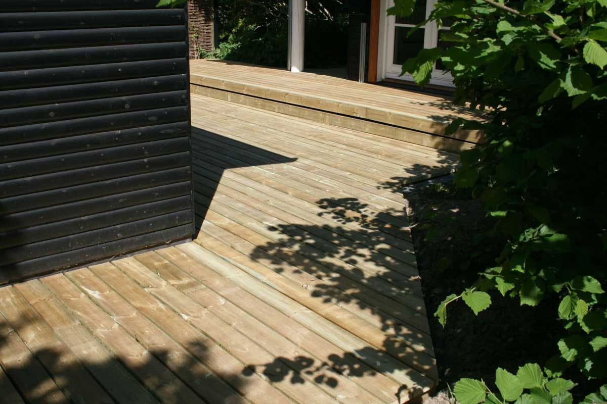 Træterrasse med vinklede planker, der støder op til en mørk lamelvæg og frodige grønne områder. Solrige udendørs omgivelser.