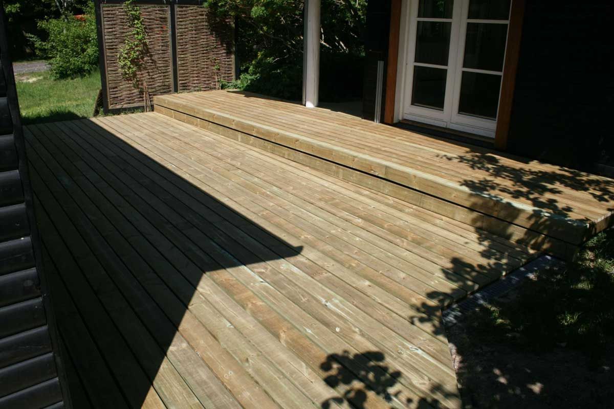 Træterrasse med trapper, der fører til en dør, foran en bygning med et flettet hegn.