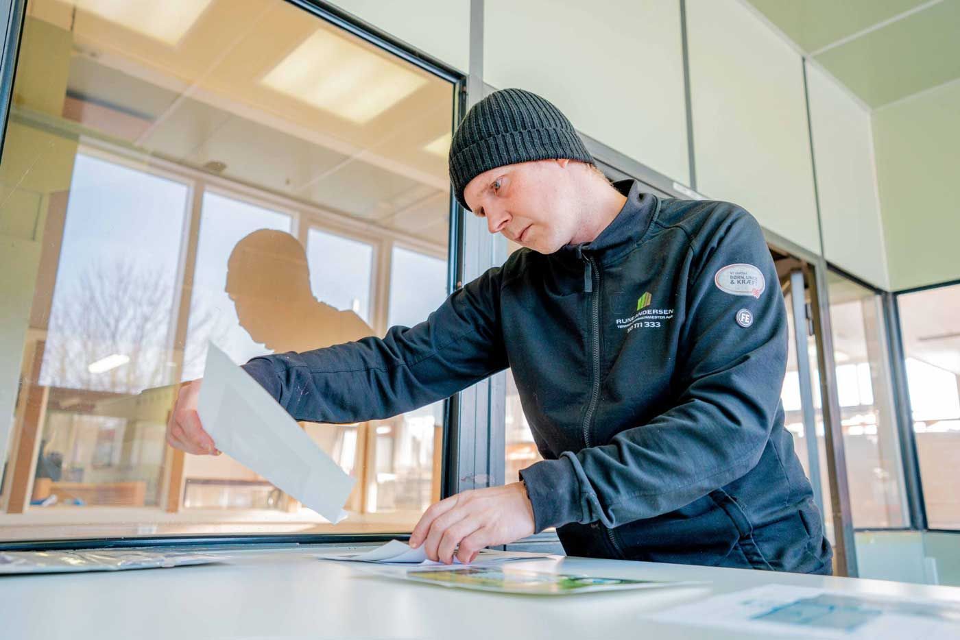 Mand i sort jakke og hue undersøger papirer ved et vindue.