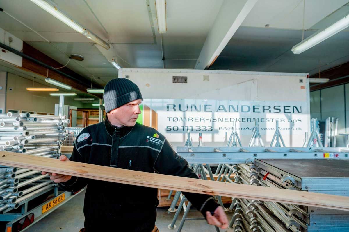 Mand i et værksted holder en træplanke, kigger til venstre. Stillads og udstyr i baggrunden.