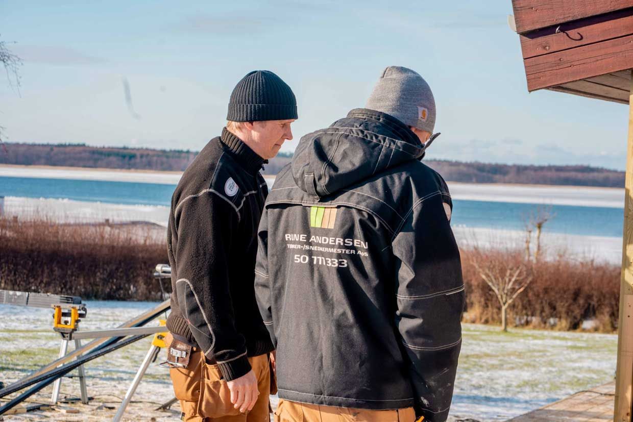 To personer i vinterjakker undersøger et tag foran et snedækket landskab og vand.