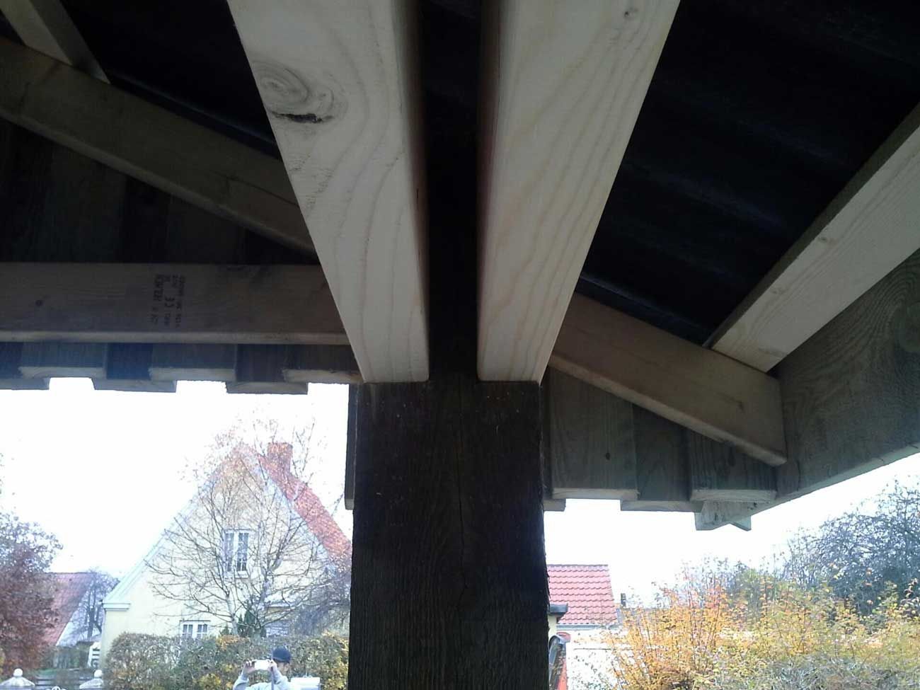 Træbjælker danner en tagkonstruktion over en mørk overflade, understøttet af en lodret stolpe.